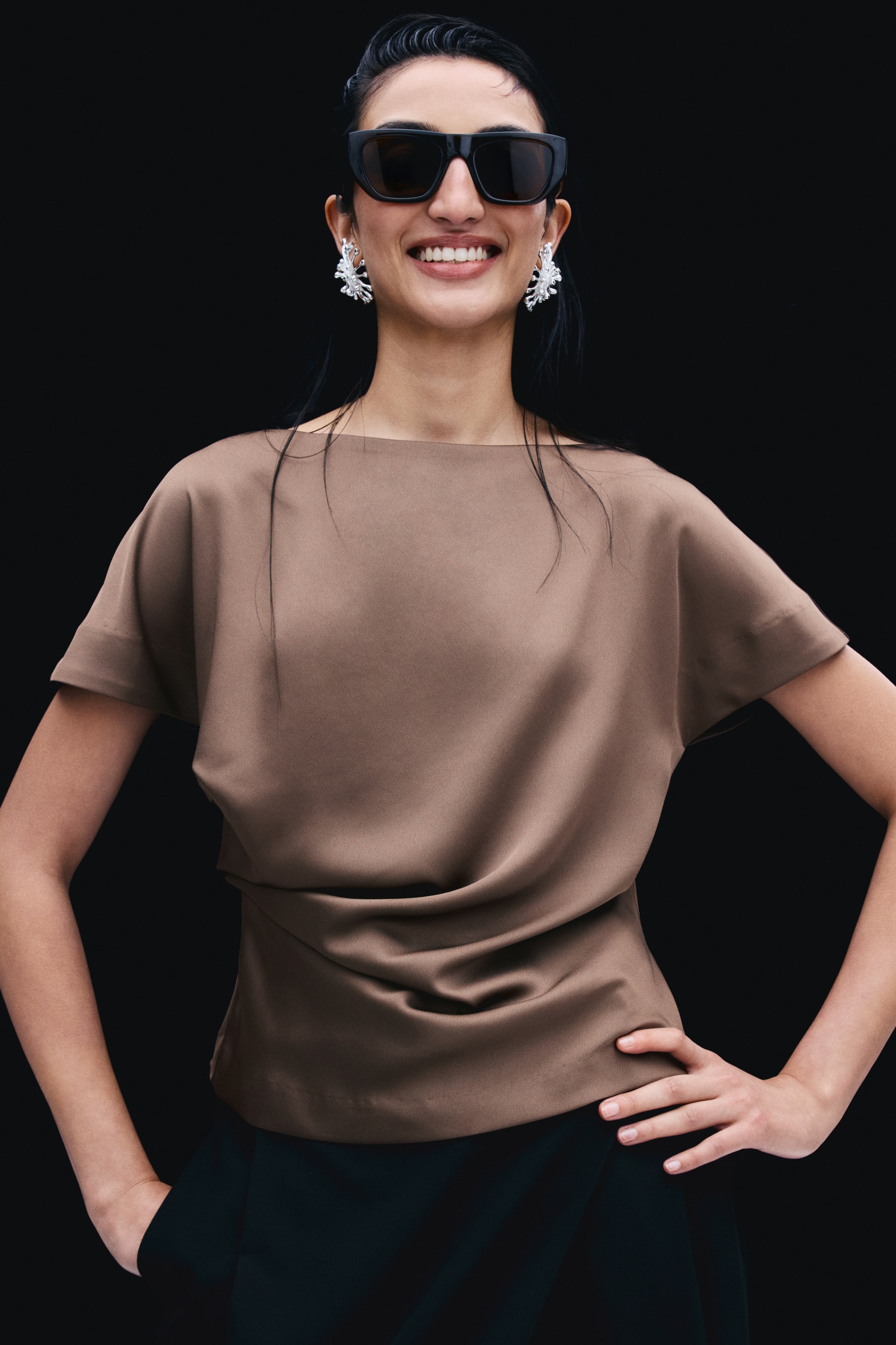 View larger image: A brown satin blouse featuring a boat neckline, cap sleeves, and draped pleating at the waist is worn by the model. The blouse is styled with black trousers, black sunglasses, and silver statement earrings.