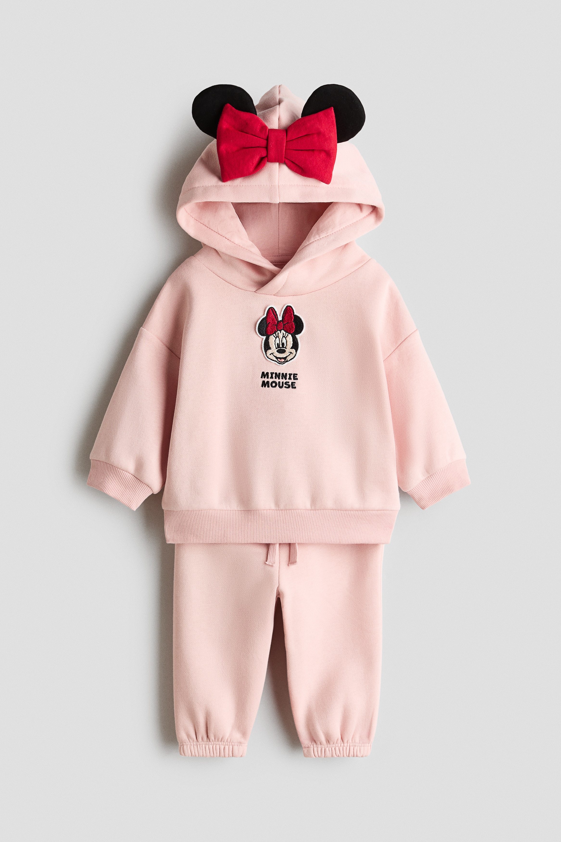 A light pink fleece hoodie and track pants set is shown from the front, featuring a hoodie with a Minnie Mouse graphic on the chest and a hood detailed with 3D ears and a red bow. The matching track pants are styled with an elasticated waistband, a decorative drawstring, and elasticated hems.
