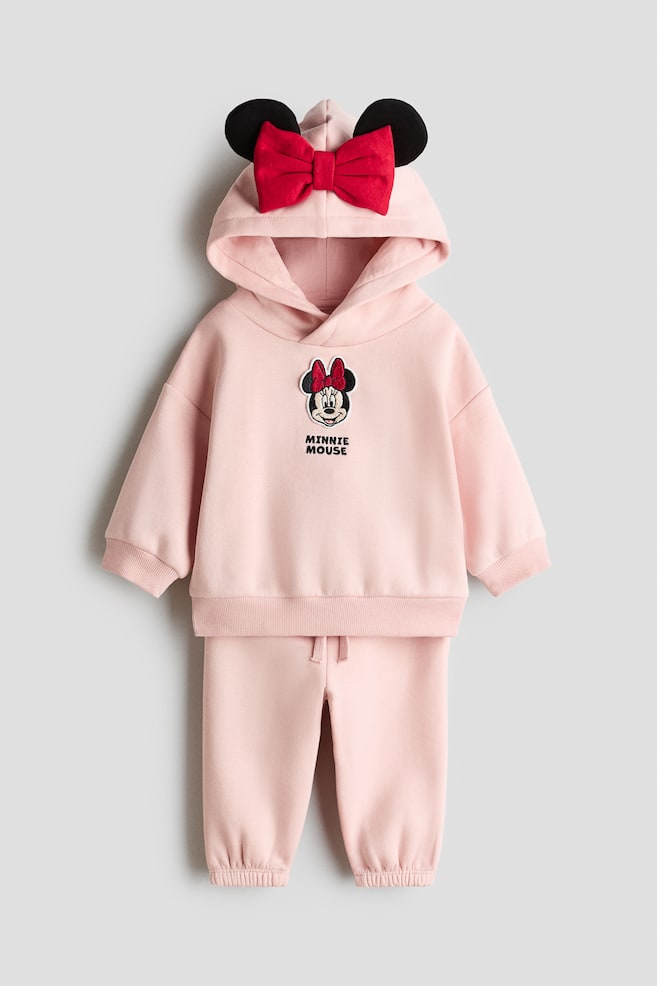 A light pink fleece hoodie and track pants set is shown from the front, featuring a wrap-front hood detailed with black 3D ears and a large red bow. The hoodie is displaying dropped shoulders, a Minnie Mouse graphic on the chest, and ribbed trims, while the matching pants are featuring an elasticated drawstring waist and elasticated hems. - 1