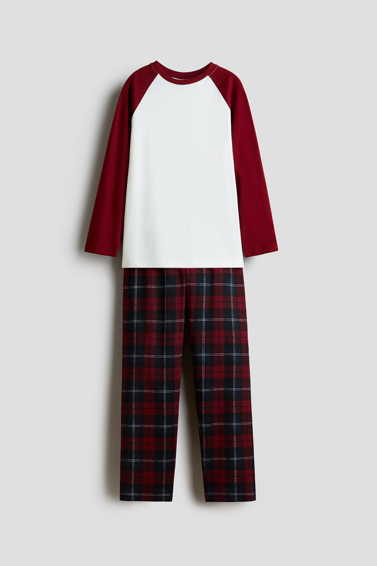 A two-piece pajama set is shown front-facing, featuring a long-sleeved raglan top with a solid white body and contrasting burgundy sleeves. The coordinating straight-leg pants feature a classic red, navy, and white plaid pattern.