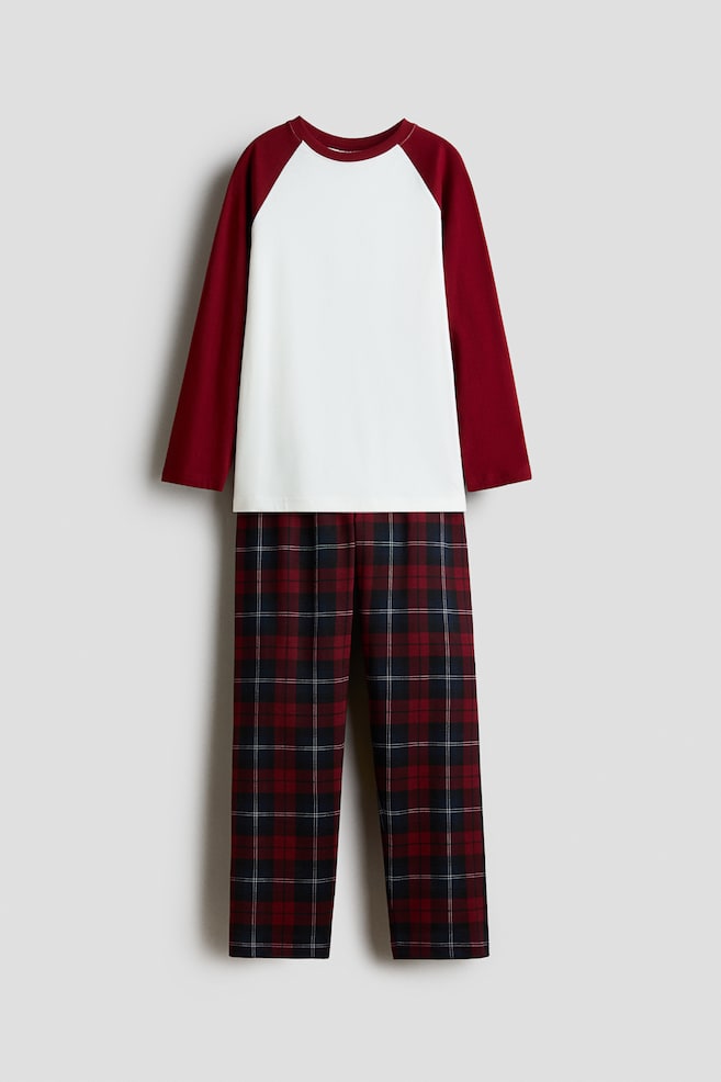 A two-piece pyjama set is shown front-facing, featuring a long-sleeved raglan top with a solid white body and contrasting burgundy sleeves. The matching straight-leg trousers feature a classic red, navy and white plaid pattern. - 1