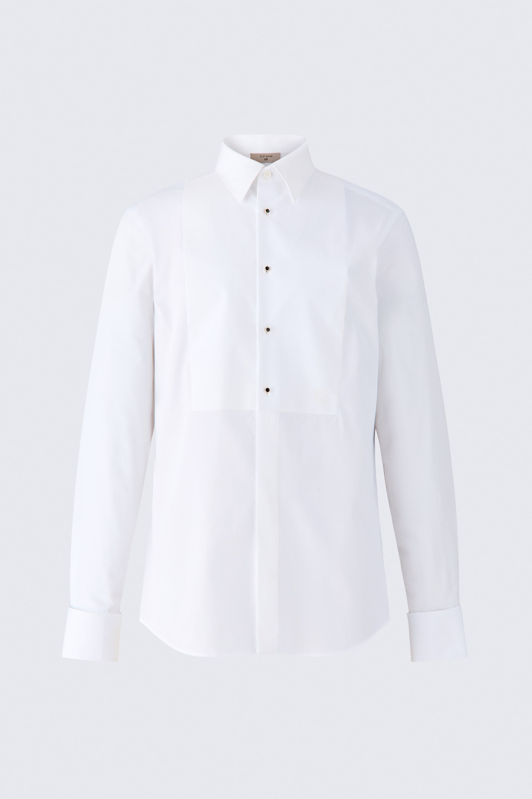 View larger image: A white cotton tuxedo shirt is shown from the front, featuring a structured bib panel on the chest with four black stud buttons. The shirt has a classic spread collar, long sleeves with French cuffs, and a concealed button placket running down the lower front.