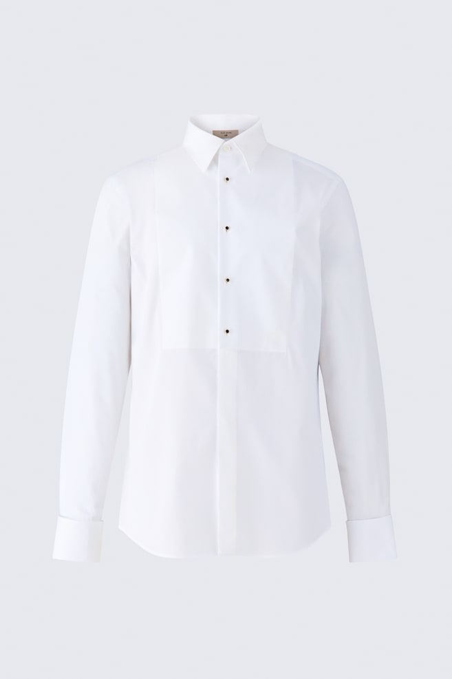 A white cotton tuxedo shirt is shown front-facing, featuring a structured bib panel on the chest with four black stud buttons. The shirt features a classic spread collar, long sleeves with French cuffs, and a concealed button placket running down the lower front. - 1