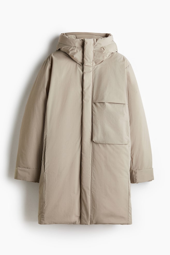 La parka Regular Fit Padded est présentée de face, dans une couleur beige clair. Elle est dotée d'une capuche attachée avec des cordons de serrage réglables, d'une fermeture frontale dissimulée sous un rabat-tempête, et d'une grande poche à rabat sur le côté droit du porteur. Des poches fendues verticales sur les côtés sont également visibles, et ses longues manches sont retroussées aux poignets. - 1