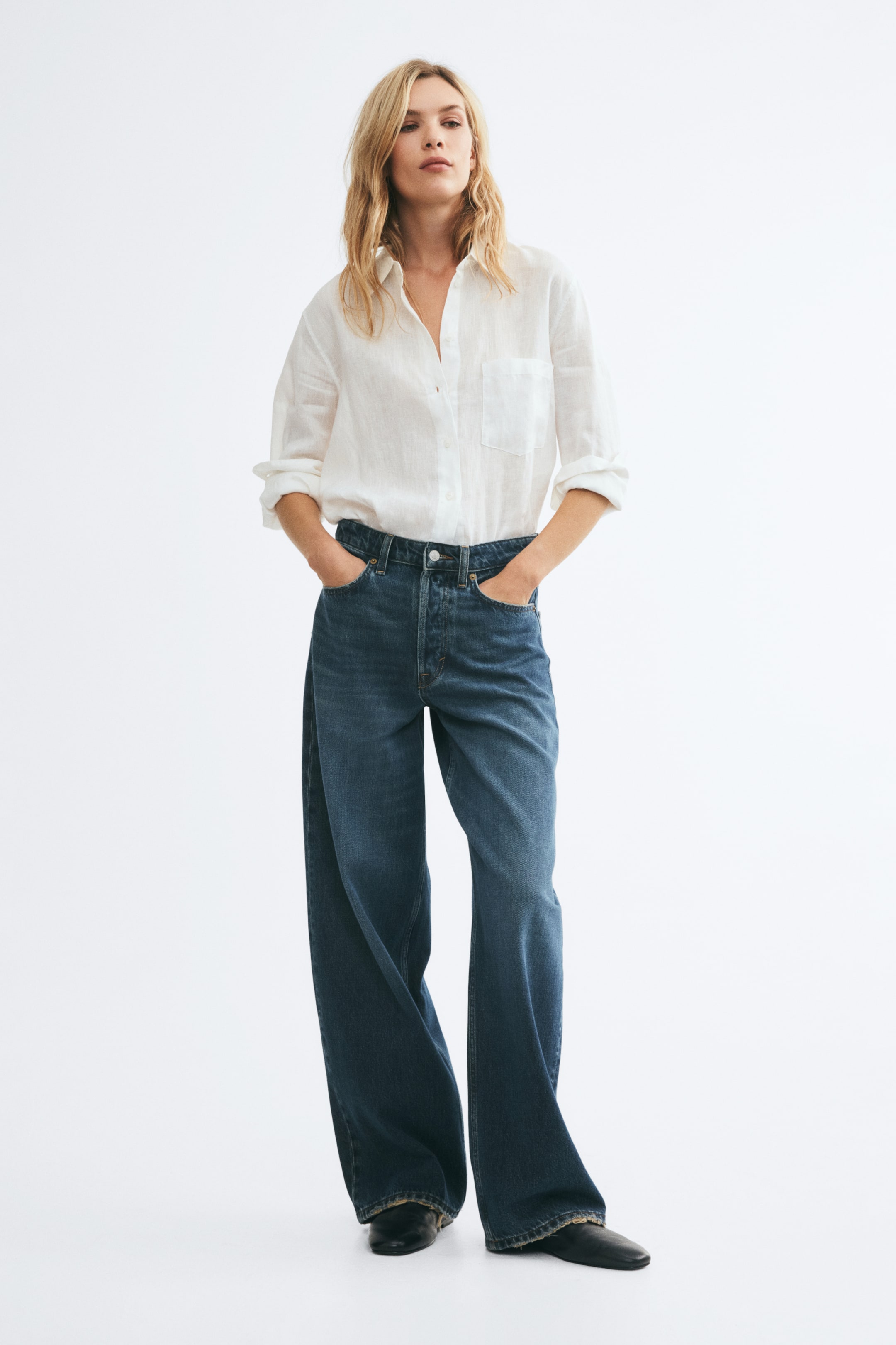 Grotere afbeelding bekijken: Blauwe wijde denim jeans met een losse, baggy pasvorm en een normale taille worden gedragen door het model, dat rechtop staat met de handen in de voorzakken. De jeans zijn gestyled met een ingestopte witte hemdblouse met knopen en zwarte schoenen.