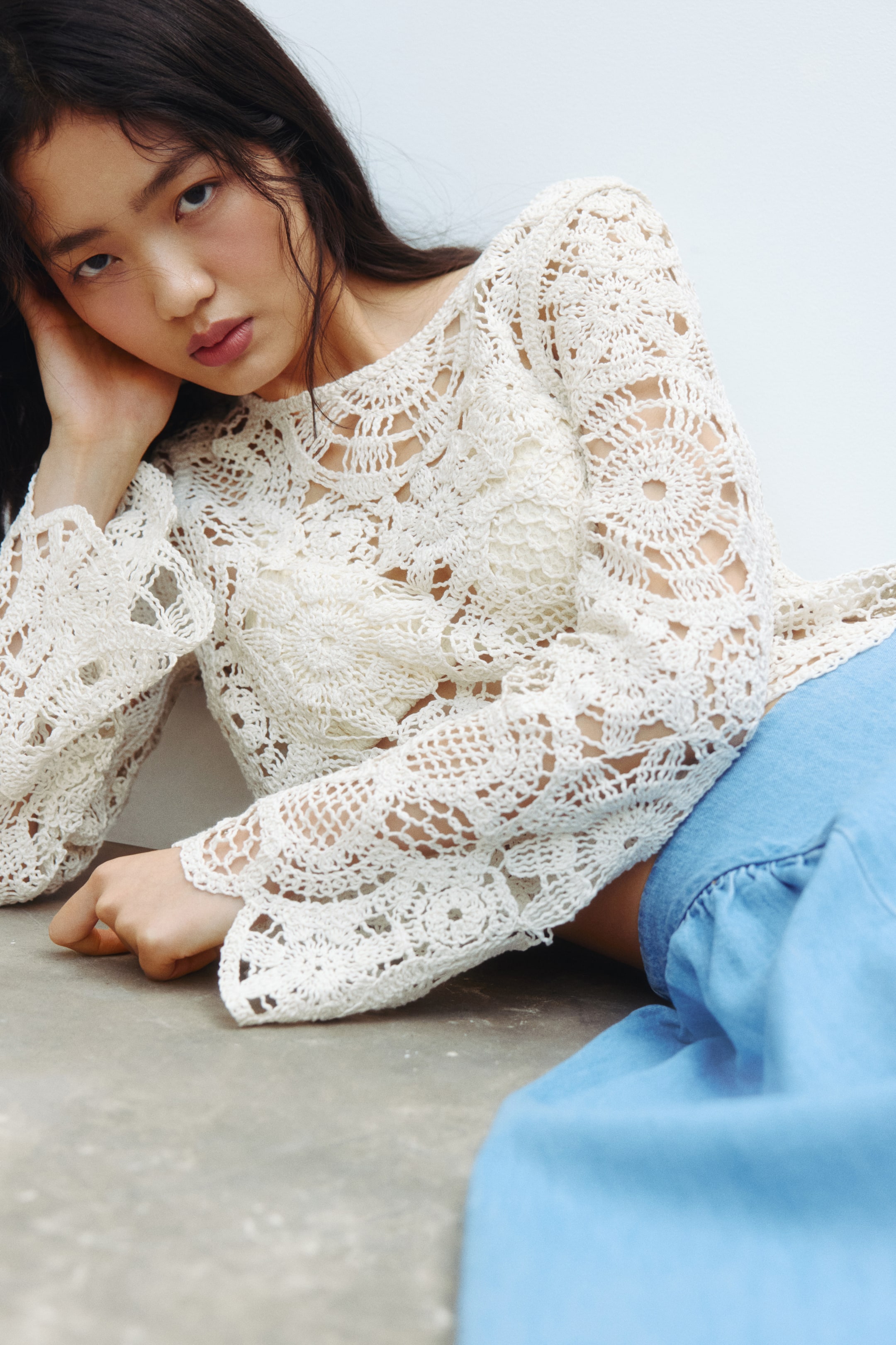 View larger image: An off-white crochet top features intricate openwork patterns with large circular motifs throughout, a wide boat neckline with a scalloped edge, and long bell sleeves. The top softly drapes over a blue garment, highlighting its textured design and relaxed fit.