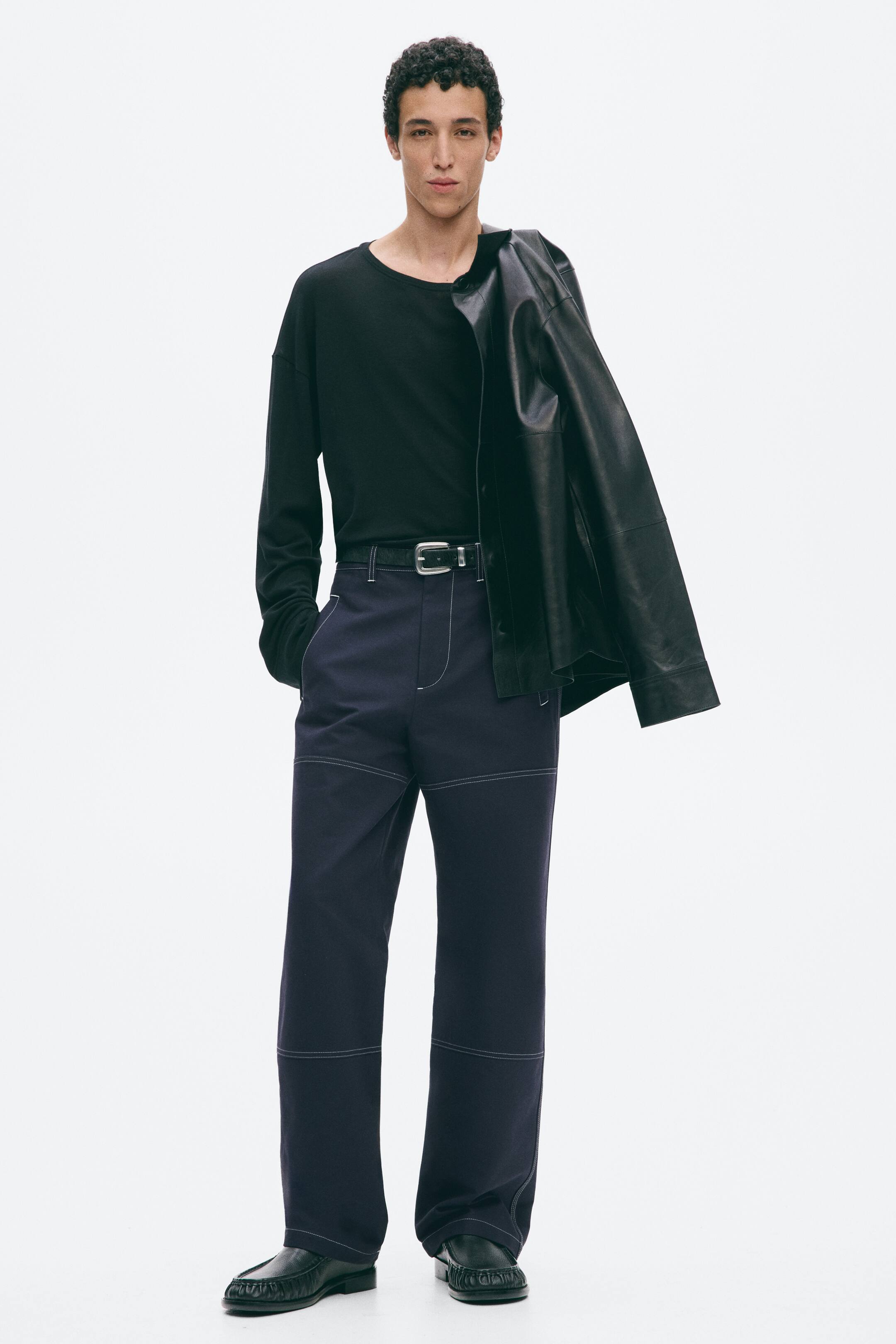 Vezi imaginea mai mare: Pantaloni bleumarin din țesătură de bumbac, cu cusături albe contrastante, vizibile pe toată suprafața, purtați de model, având închidere cu fermoar și nasture, buzunare oblice cu refilet și croială dreaptă. Pantalonii, concepuți cu o croială lejeră, sunt prinși cu o curea neagră, iar mâna stângă a modelului se odihnește într-un buzunar din față.
