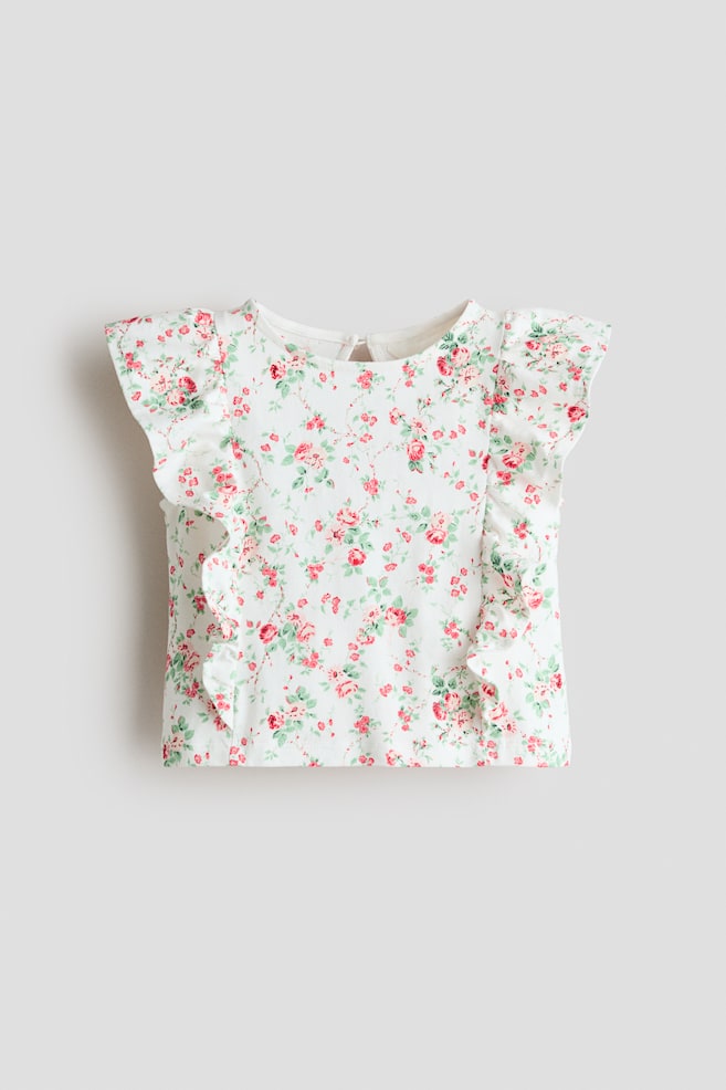 A white top is facing front, featuring a floral pattern of pink roses and green leaves. It has short, ruffled cap sleeves, a round neckline, and vertical ruffles on the front bodice, with a keyhole closure and button visible at the back of the neck. - 1
