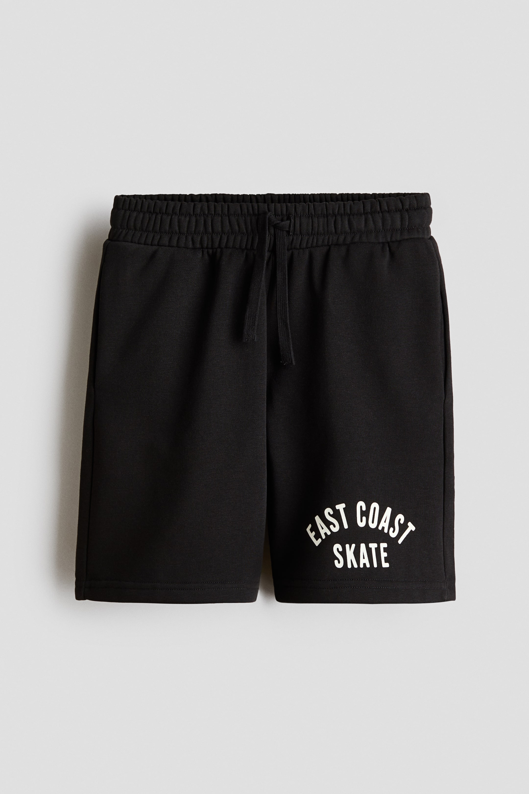 Größeres Bild anzeigen: Knielange, locker sitzende Shorts erscheinen in schwarzem Sweatshirt-Stoff mit Print und präsentieren ihre Vorderansicht. Sie verfügen über einen elastischen Kordelzugbund und einen weißen "EAST COAST SKATE"-Aufdruck am unteren rechten Bein.