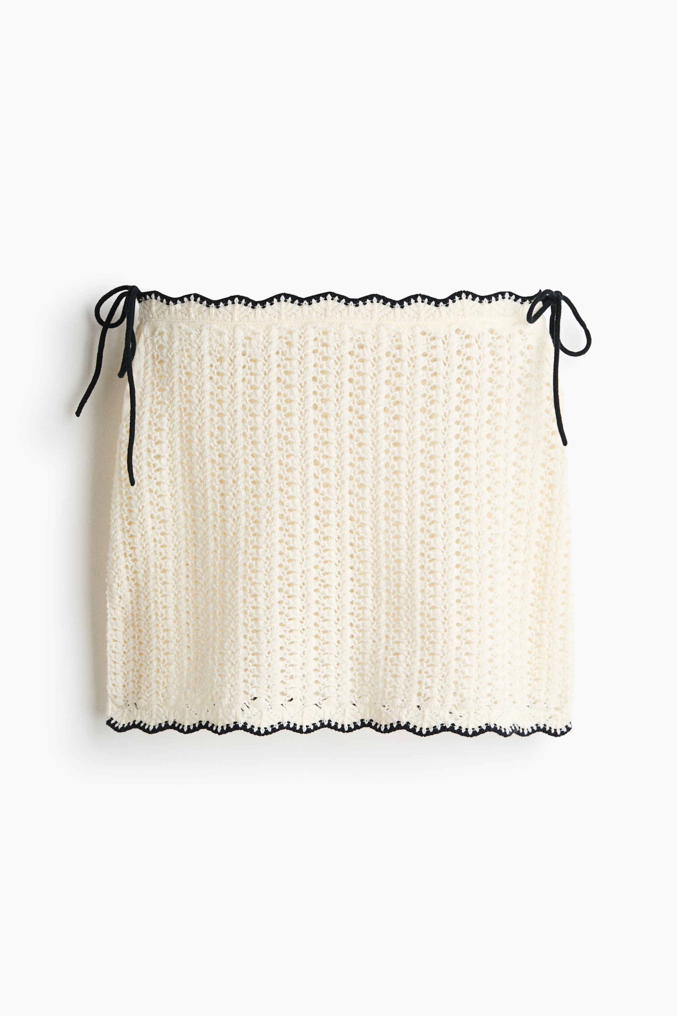 Grotere afbeelding bekijken: Een aansluitende minirok presenteert zijn voorkant, gemaakt van een off-white breisel met een haakwerk-look en zwarte geschulpte randen langs de boven- en onderzoom, met smalle zwarte strikjes die aan elke kant van de bovenkant zijn geknoopt.