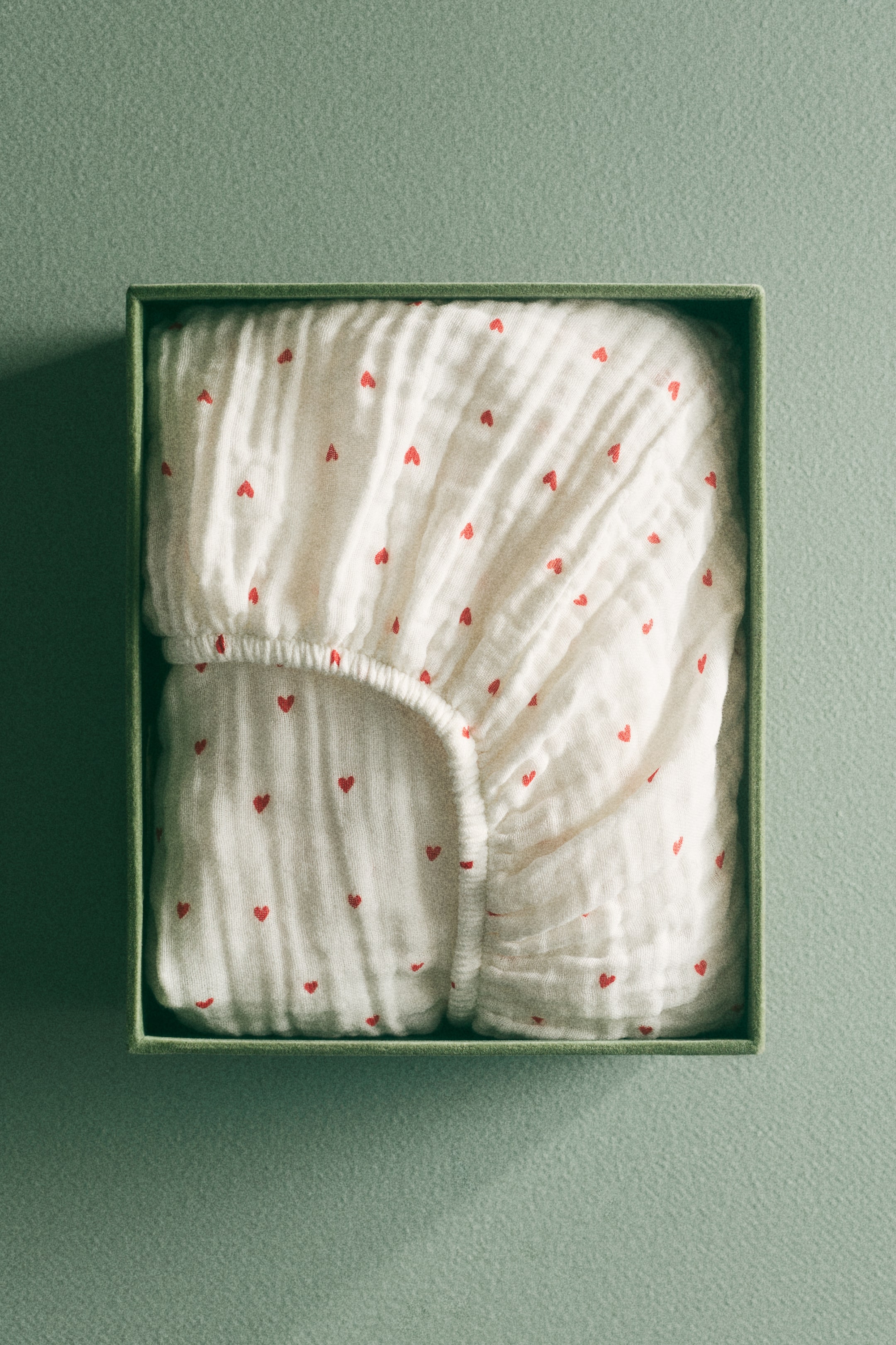 拡大画像を表示: フィットシーツは折りたたまれ、緑色の四角い箱の中で表面が見えています。このシーツは柔らかいコットンモスリン製で、小さな赤いハートの総柄が特徴です。伸縮性のある縁が見えています。