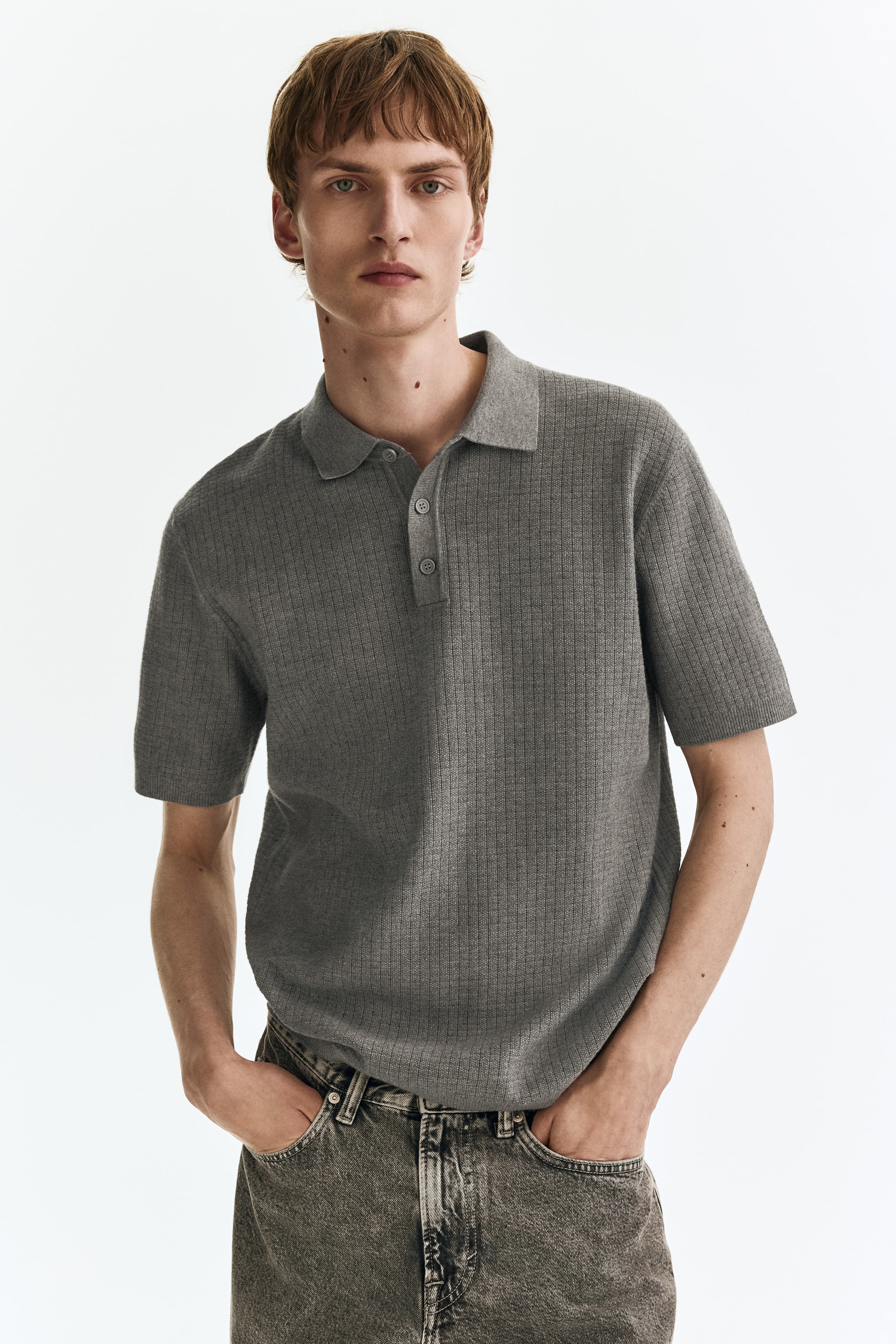 View larger image: A grey ribbed knit polo shirt is being worn, facing forward, featuring a classic polo collar, short sleeves, and a three-button placket with matching grey buttons.