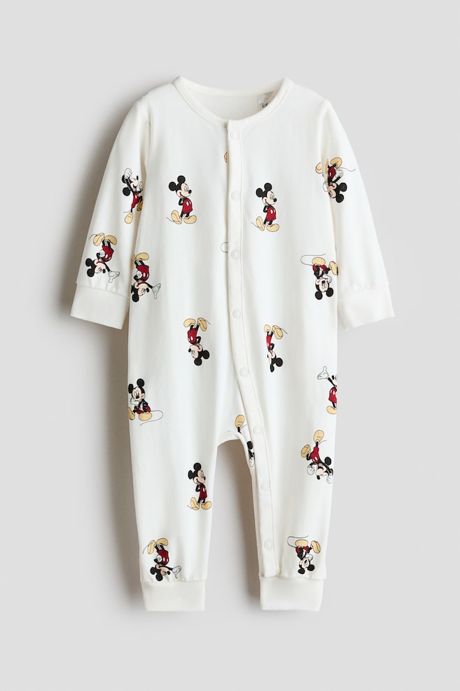 Vista frontal de un pijama enterizo de algodÃ³n jersey en color blanco roto, de manga larga, con un diseÃ±o estampado integral de personajes de Mickey Mouse. El pijama enterizo presenta cuello redondo, cierre con botones a presiÃ³n que se extiende por el delantero y a lo largo de una pernera, y amplios acabados de canalÃ© en los puÃ±os y los bajos. - 1