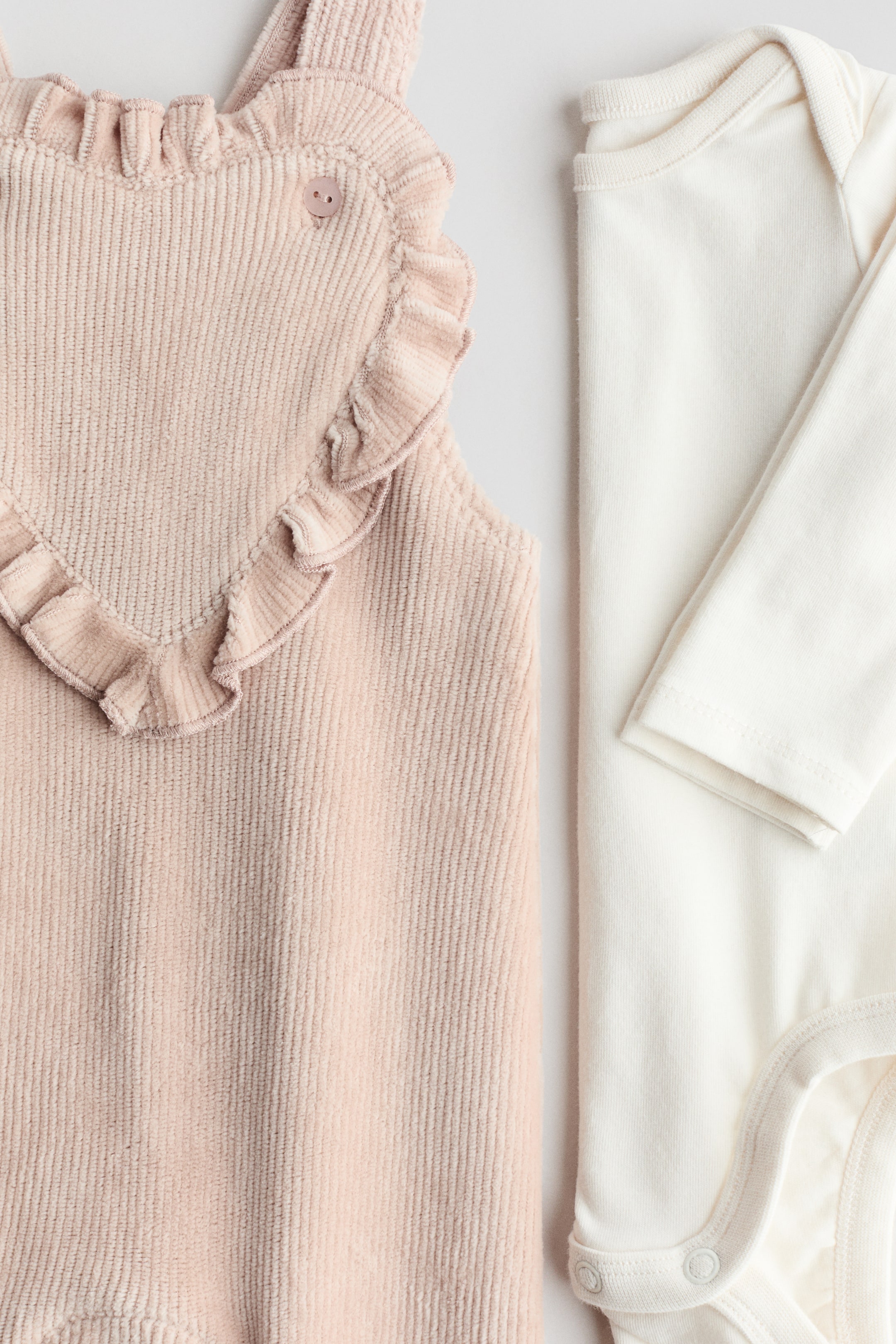 View larger image: A pink corduroy overall with ruffled, heart-shaped bib front and a small button on a shoulder strap is lying next to a cream long-sleeved bodysuit. The overall is showcasing vertical corduroy ridges, while the bodysuit is featuring a smooth knit fabric, a ribbed crew neckline, and snaps at the crotch.