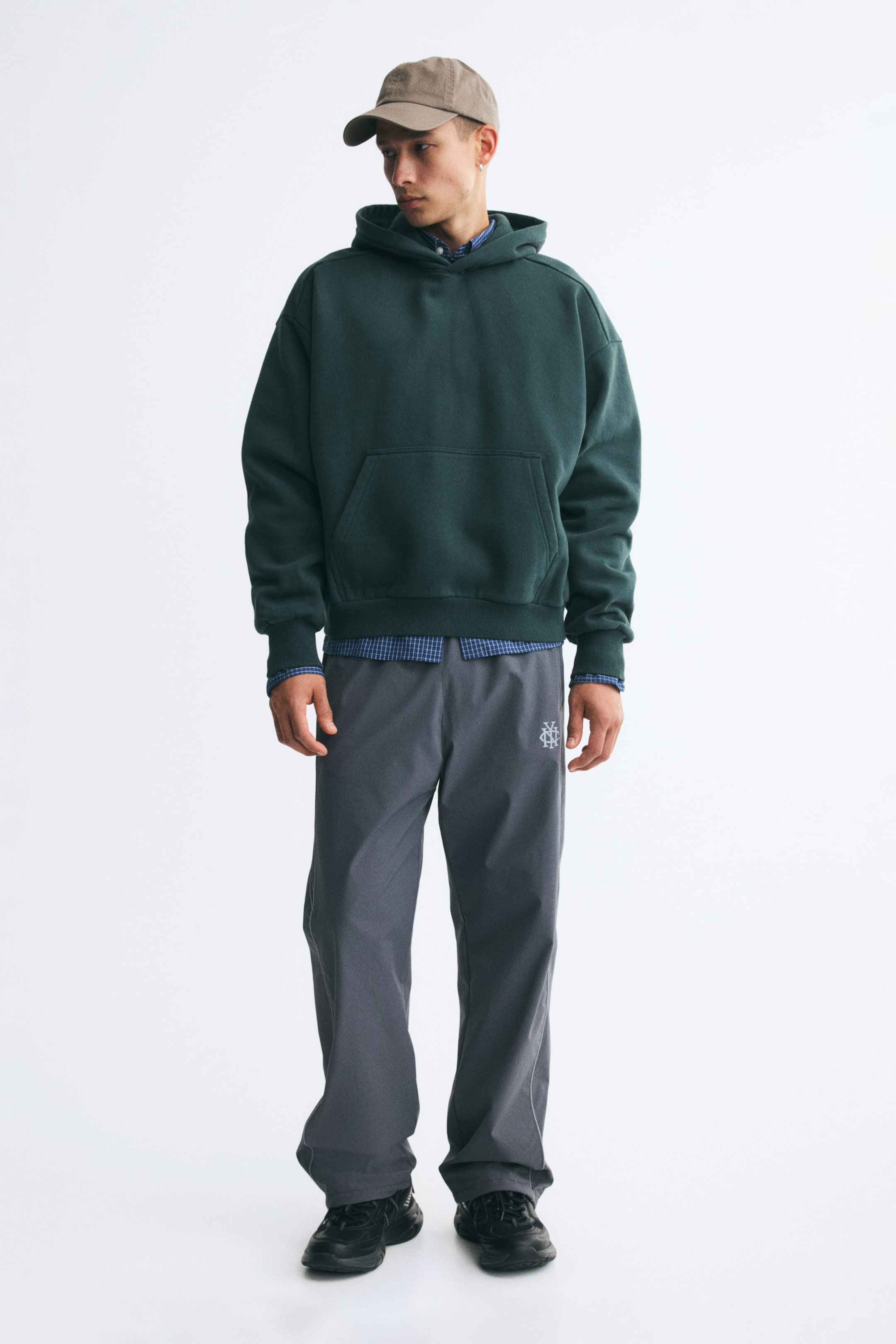 Grotere afbeelding bekijken: Een donkergroene, oversized hoodie wordt gedragen door het model en is voorzien van verlaagde schoudernaden, een kangoeroezak en geribde boorden aan de mouwen en zoom. De hoodie wordt gecombineerd met een blauw shirt met patroon en knoopsluiting, en draagt bij aan een look met een brede grijze broek en een beige pet.
