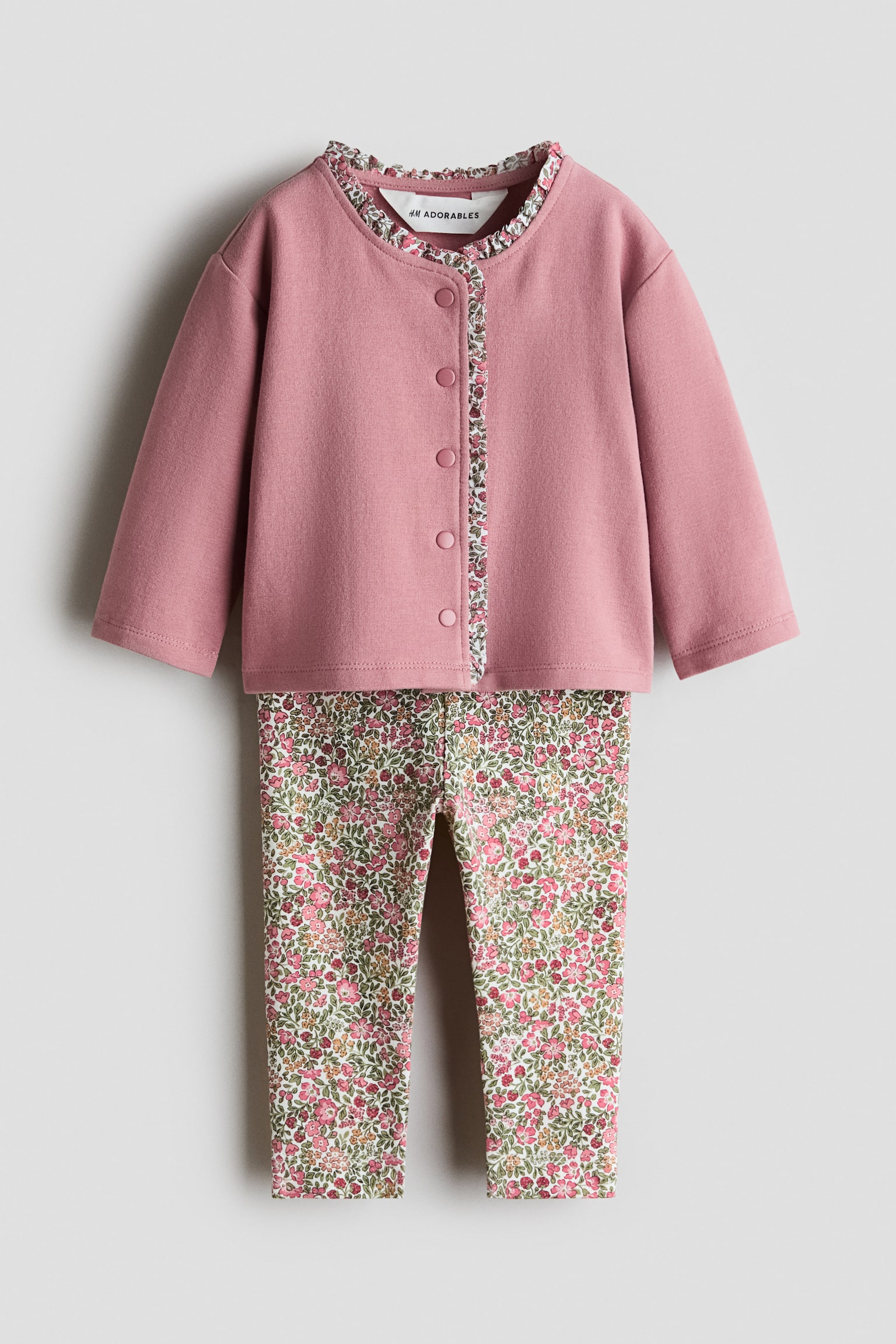 Un completo a due pezzi per bebÃ¨ Ã¨ mostrato frontalmente, composto da un cardigan rosa cipria e da leggings coordinati con stampa floreale. Il cardigan a maniche lunghe presenta una chiusura frontale con bottoni a pressione e un bordo increspato con motivo floreale lungo il colletto e lâabbottonatura interna, mentre i leggings mostrano una stampa floreale all-over in rosa, verde e bianco.