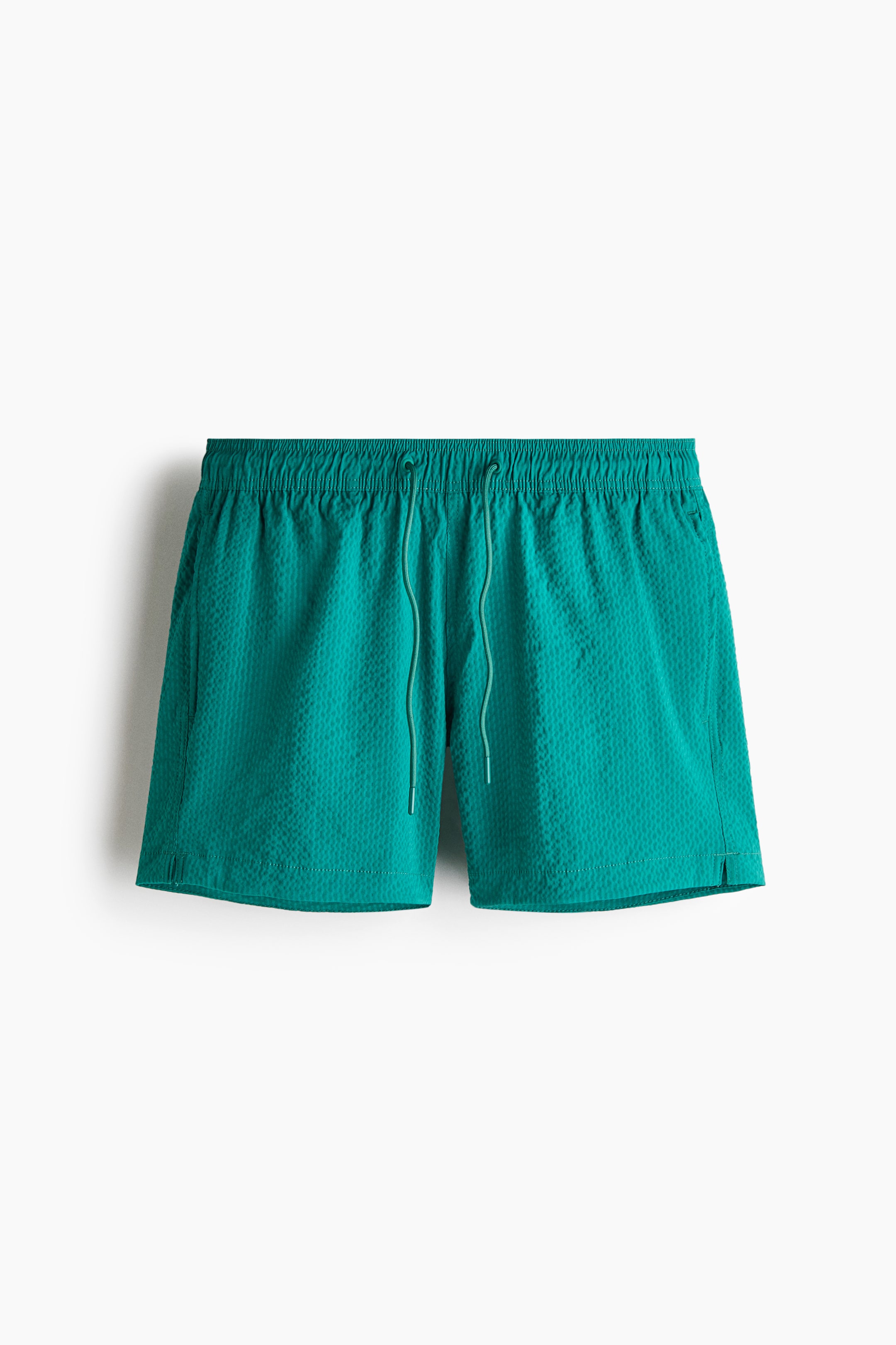 View larger image: Teal seersucker swim shorts are shown from the front, featuring a covered elasticated waistband with a visible drawstring. The shorts have vertical side pockets and small slits on each side of the hem.