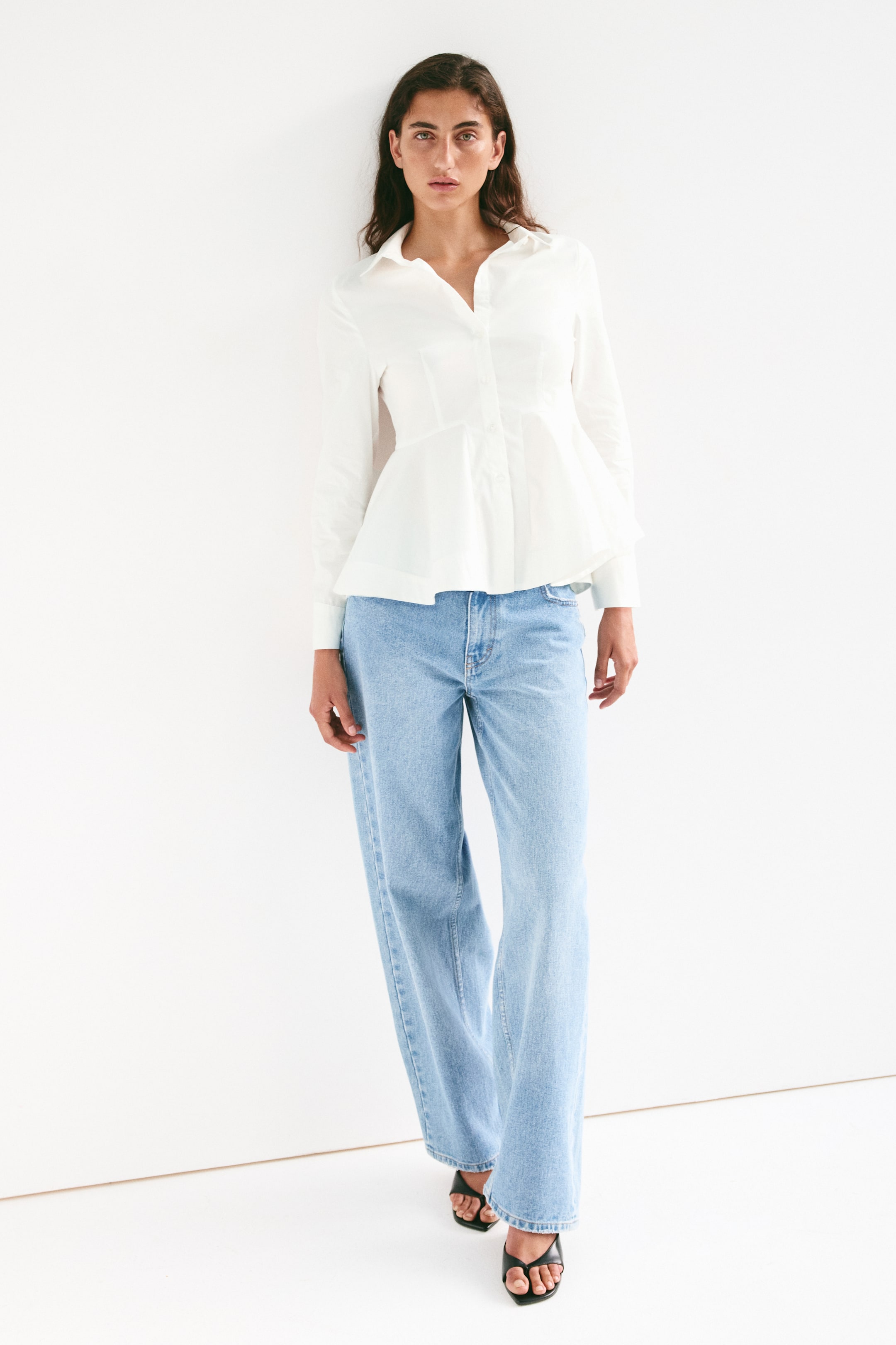 View larger image: The model is wearing a white fitted shirt in an airy cotton weave. It features a collar, front button closure, a yoke at the back, and long sleeves with buttoned cuffs. The shirt is designed with a visible seam at the waist, with a flared peplum extending below, paired with light-wash jeans.