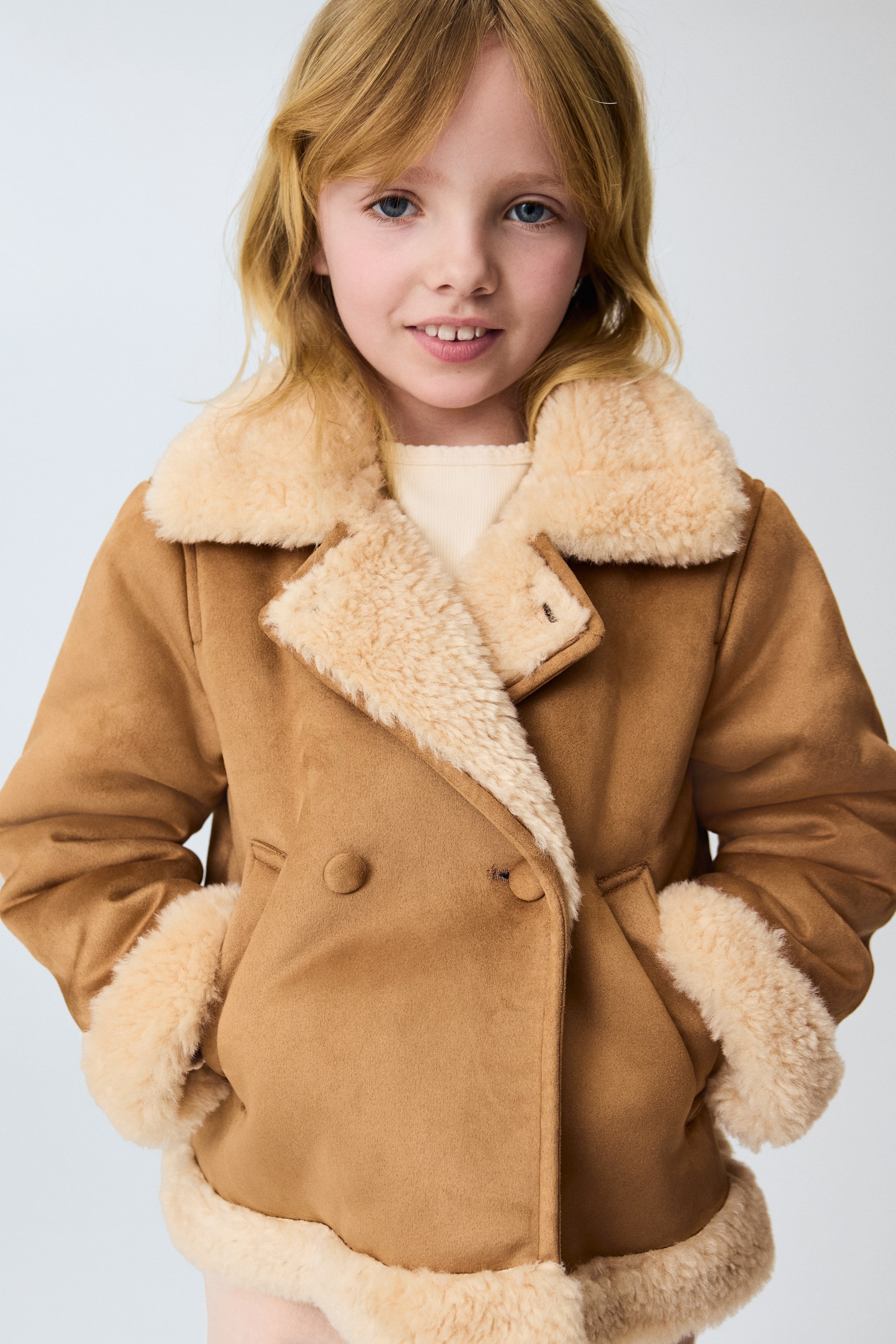 View larger image: A brown faux suede coat featuring wide notched lapels, cuffs, and hem, all trimmed with cream faux shearling. Designed with a double-breasted front, showcasing two visible buttons and slanted pockets, and worn over a light-coloured ribbed top.