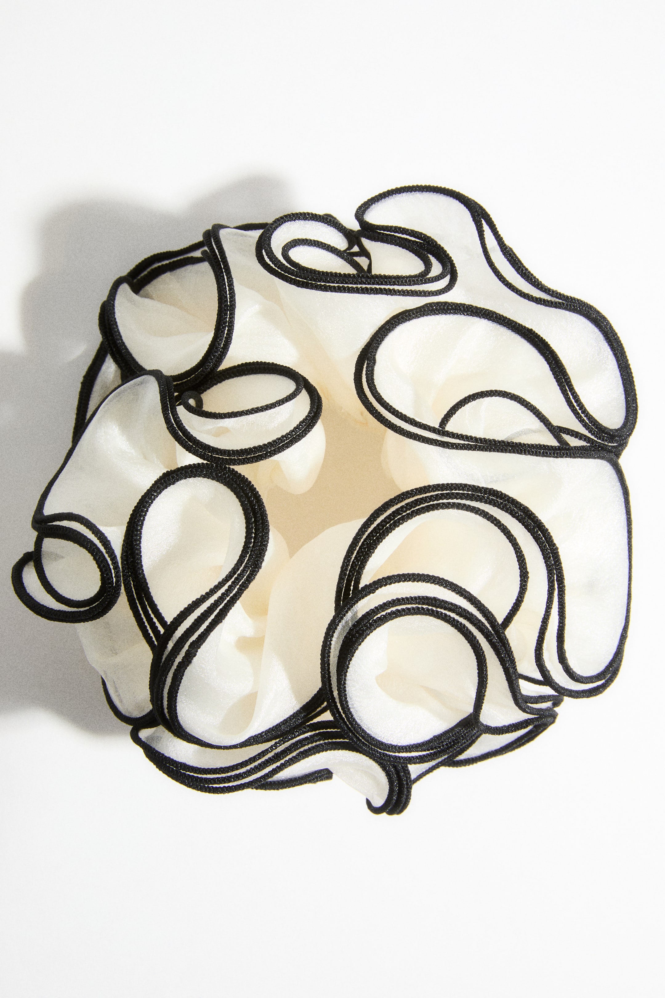 이미지 확대 보기: 크림색 패브릭 액세서리가 위에서 아래로 내려다보는 각도로 제시되어 있으며, 볼륨감 있는 주름진 형태를 보여줍니다. 검은색 트림이 얇은 패브릭 주름의 가장자리를 따라 윤곽을 형성하며, 동심원의 물결 무늬를 이룹니다.