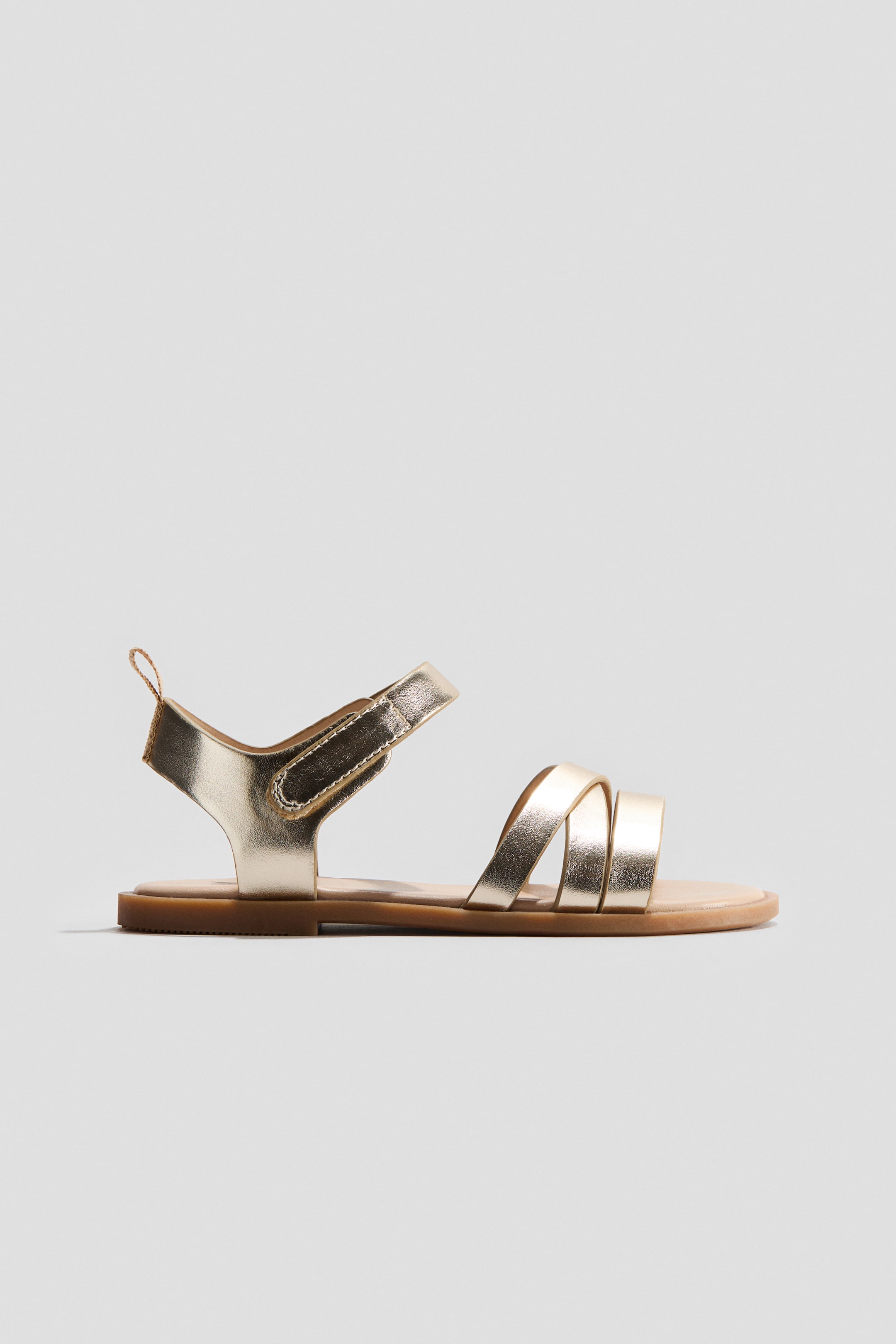 View larger image: "A golden sandal is showcased in a side view, adorned with three criss-cross straps across the foot. It features an adjustable ankle strap with a hook and loop closure, set on a flat, light brown sole."