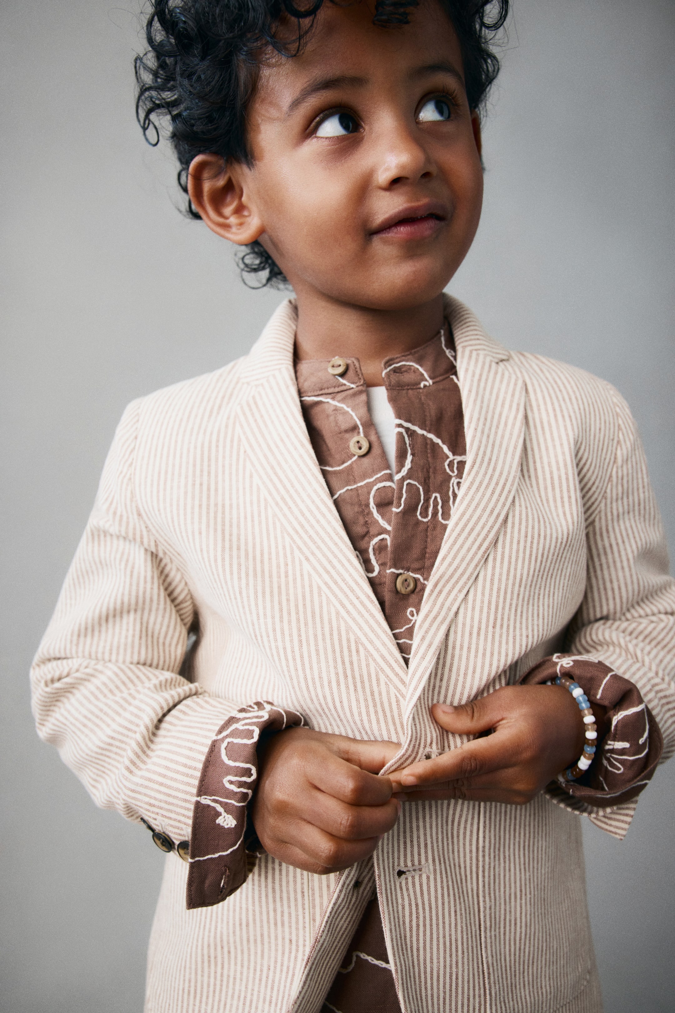 View larger image: A front view of a jacket in a striped beige and brown linen and cotton weave, featuring notch lapels and a button closure. The jacket is being worn over a brown patterned shirt, with the long sleeves rolled up at the cuffs to reveal the shirt underneath.