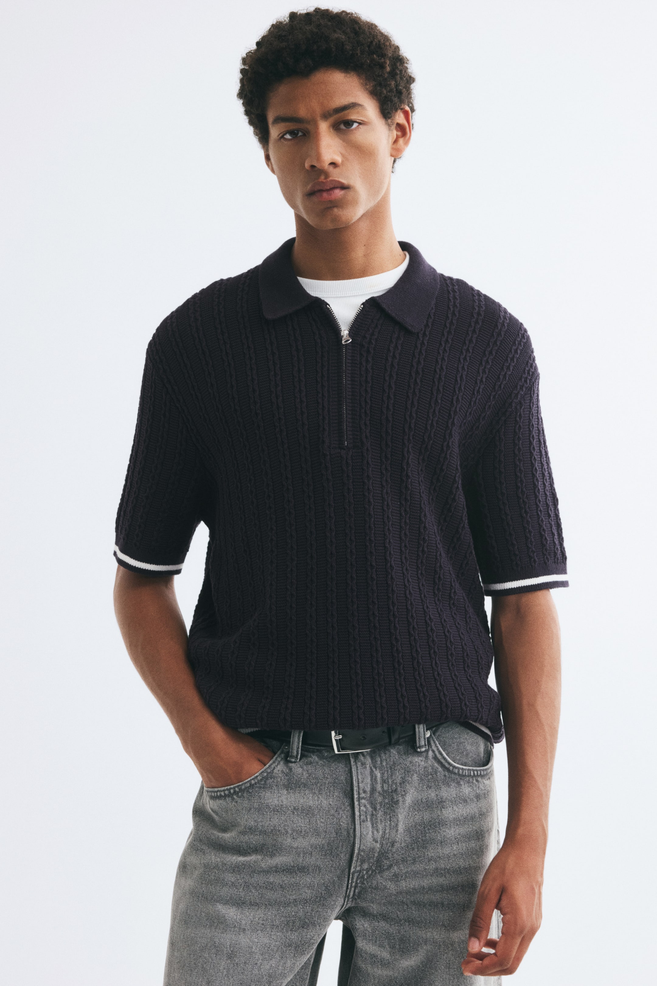 View larger image: A dark-coloured textured knit polo shirt featuring a half-zip closure, a ribbed collar, and short sleeves with white-tipped cuffs is being worn by the model. The relaxed-fit polo is layered over a white crew-neck T-shirt and styled with grey jeans.