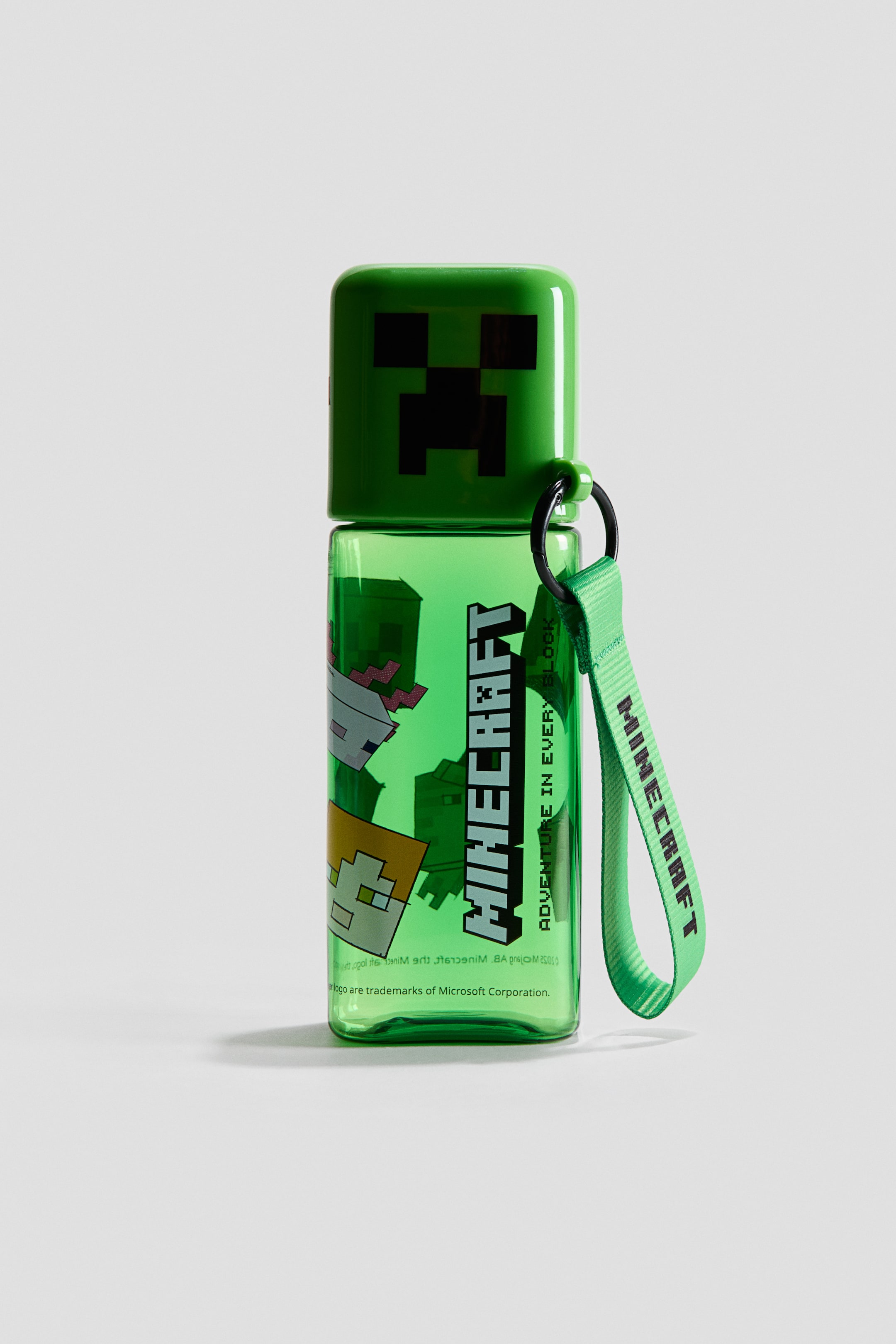 Ampliar la imagen: Una botella de agua presenta un cuerpo cilíndrico transparente de color verde con una tapa cuadrada opaca de color verde. La tapa muestra una cara de Creeper pixelada en negro, mientras que el cuerpo exhibe un logotipo vertical de 'Minecraft' y varios personajes pixelados, con una correa de tela verde que lleva el texto 'Minecraft' y está unida a la tapa.