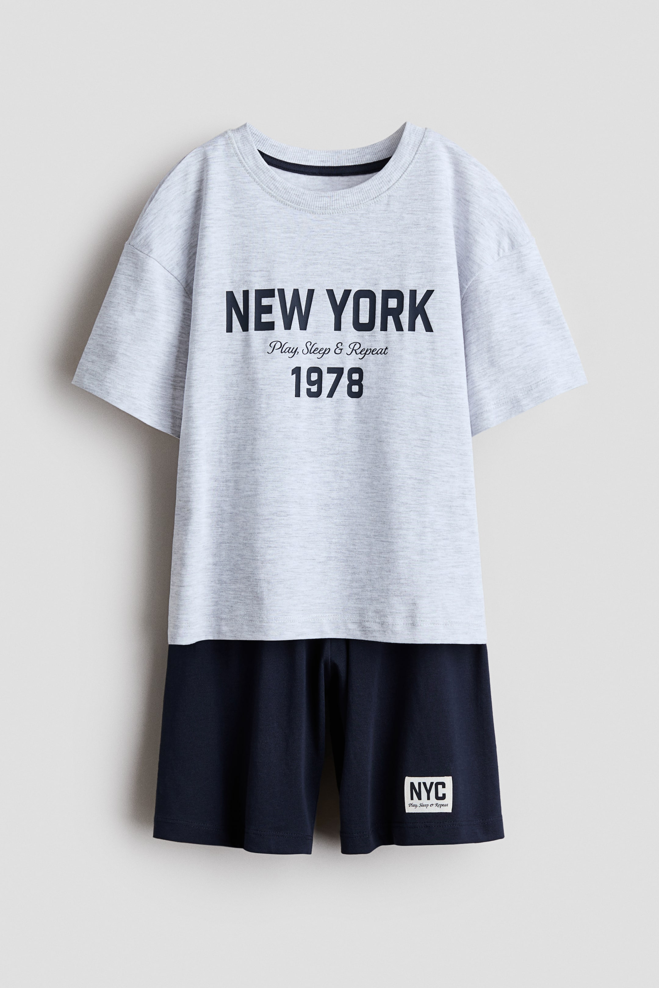 Größeres Bild anzeigen: Das Pyjama-Set zeigt seine Vorderansicht und besteht aus einem hellgrauen Marl-T-Shirt mit rundem, geripptem Halsausschnitt, überschnittenen Schultern und dem Aufdruck "NEW YORK Play, Sleep & Repeat 1978" auf der Vorderseite. Die dazugehörigen knielangen, marineblauen Shorts verfügen über einen elastischen Bund und ein weißes "NYC Play, Sleep or Repeat" Etikett am linken Bein.