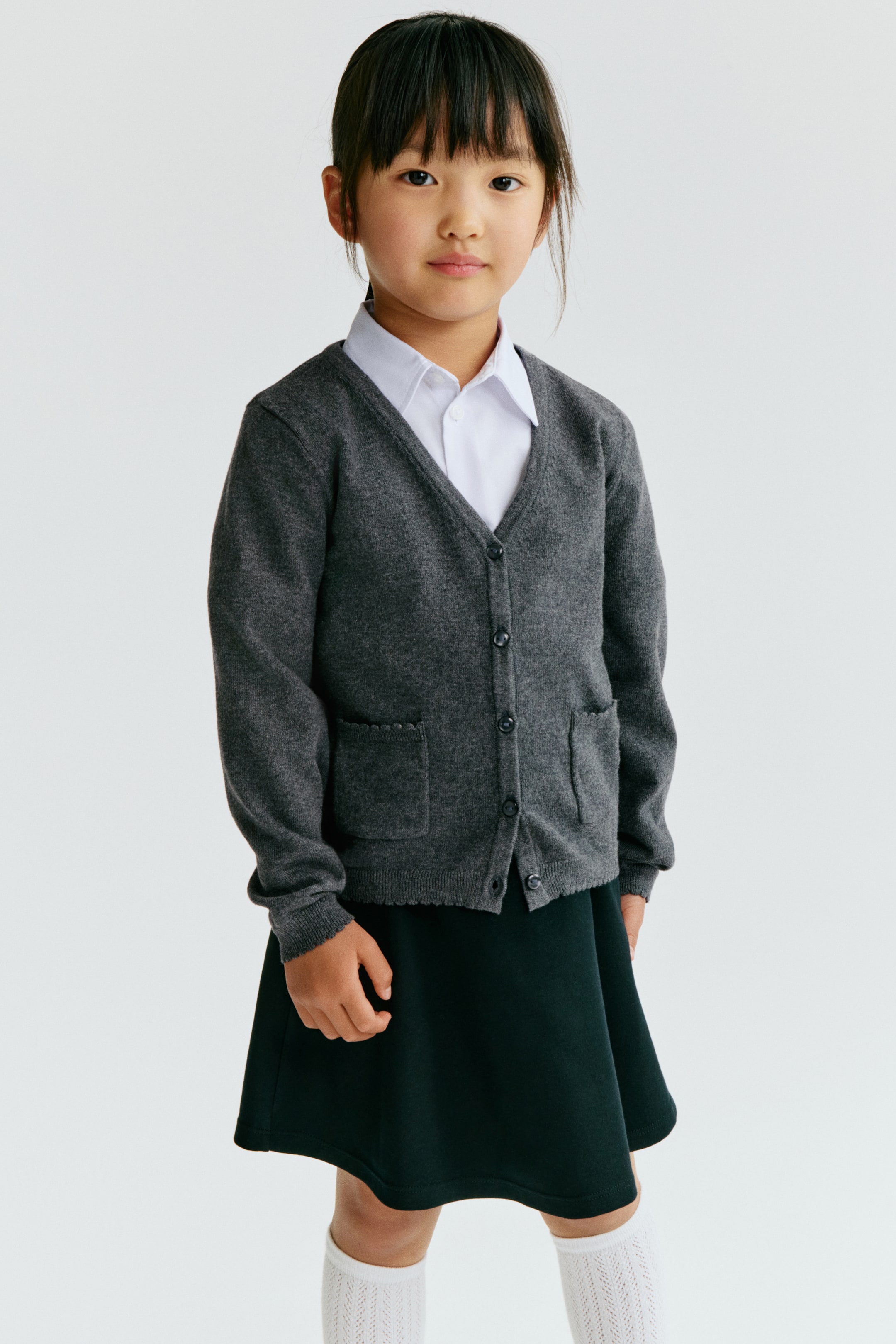 View larger image: A charcoal grey cardigan featuring a V-neckline and a button-front closure with five dark buttons. It showcases two patch pockets near the hem, with the viewer's left pocket opening having a scalloped edge, layered over a white collared shirt. Ribbed cuffs and a ribbed hem complete its design.