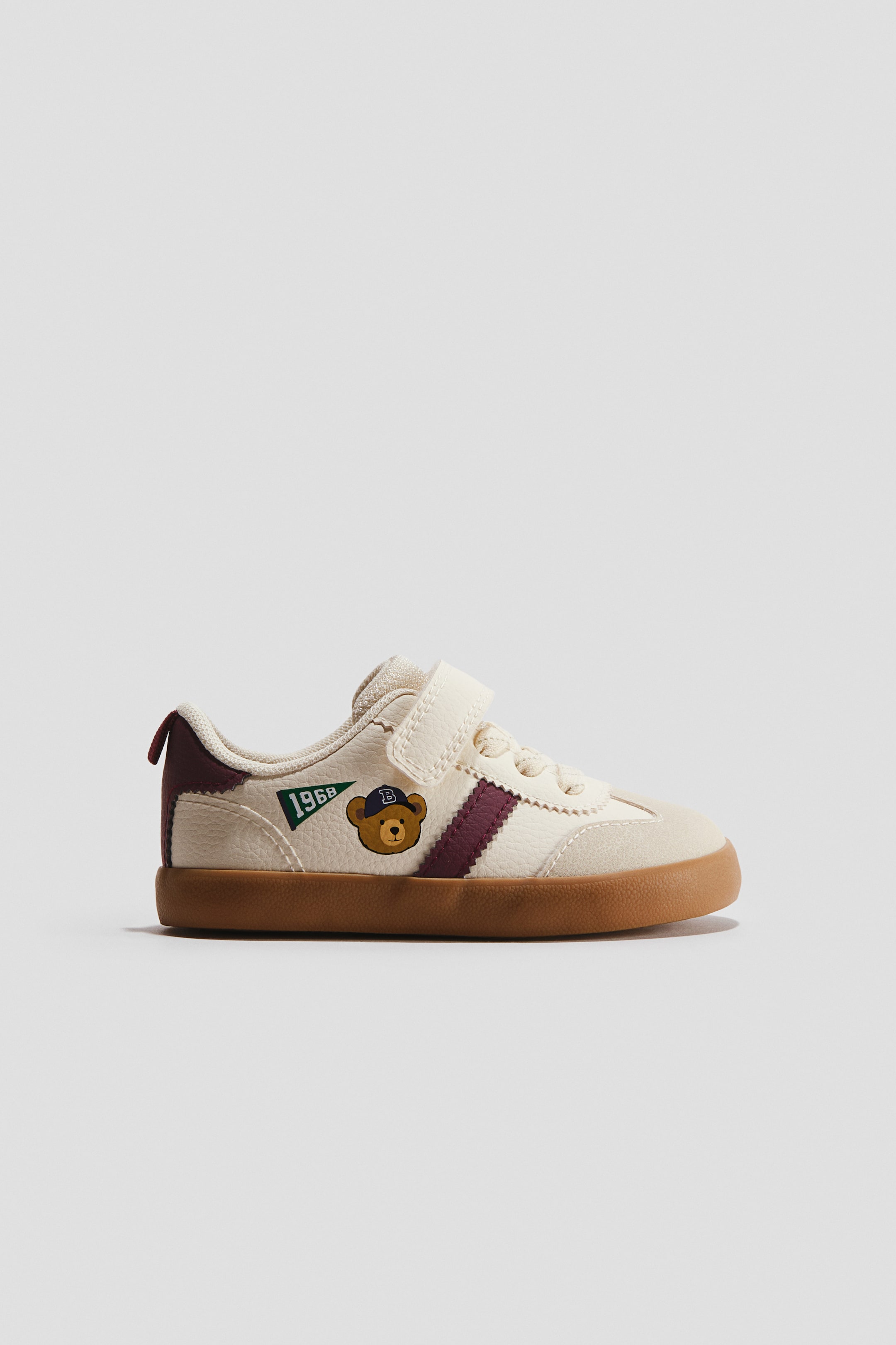 Ver imagen más grande: Zapatillas descalzas. El zapato de color crema está orientado hacia la izquierda, con una parte superior texturizada que presenta dos rayas burdeos, una cabeza de oso marrón bordada con una gorra azul con una 'B', y un banderín verde con '1968' en su lateral. Un único cierre de velcro asegura el zapato, que se completa con una puntera de imitación de ante, una lengüeta burdeos en el talón y una suela de goma color tostado.