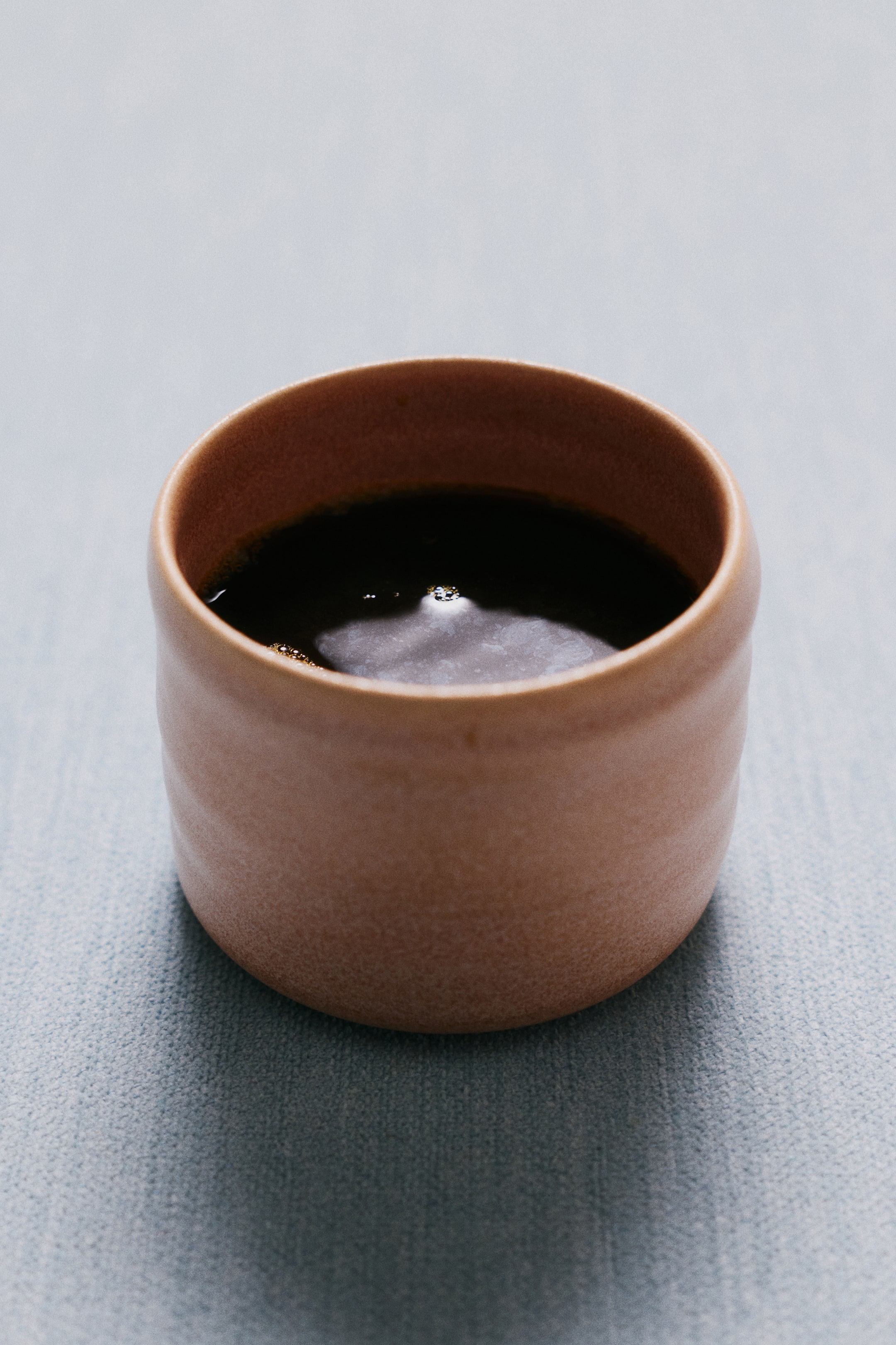 View larger image: A handleless stoneware cup with an organic shape and subtle horizontal ridges is showing a matte, earthy pink reactive glaze. The cup holds a dark liquid, highlighting its rounded rim and unique variegated texture from a slightly elevated front angle.