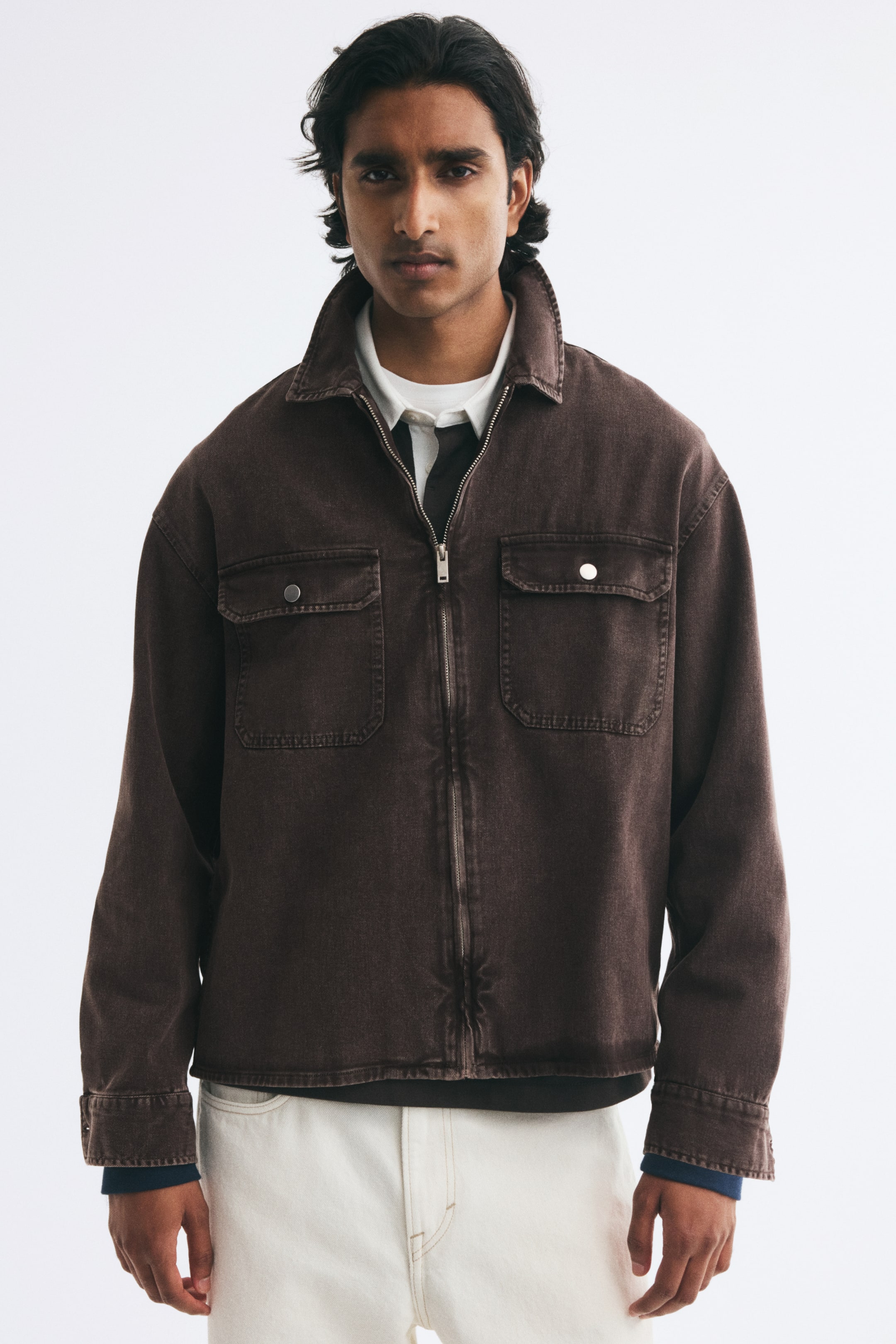 View larger image: The model is wearing a dark brown cotton twill overshirt layered over a white collared shirt and light-coloured trousers. The overshirt features a classic collar, a partially zipped front, and two patch chest pockets with buttoned flaps.