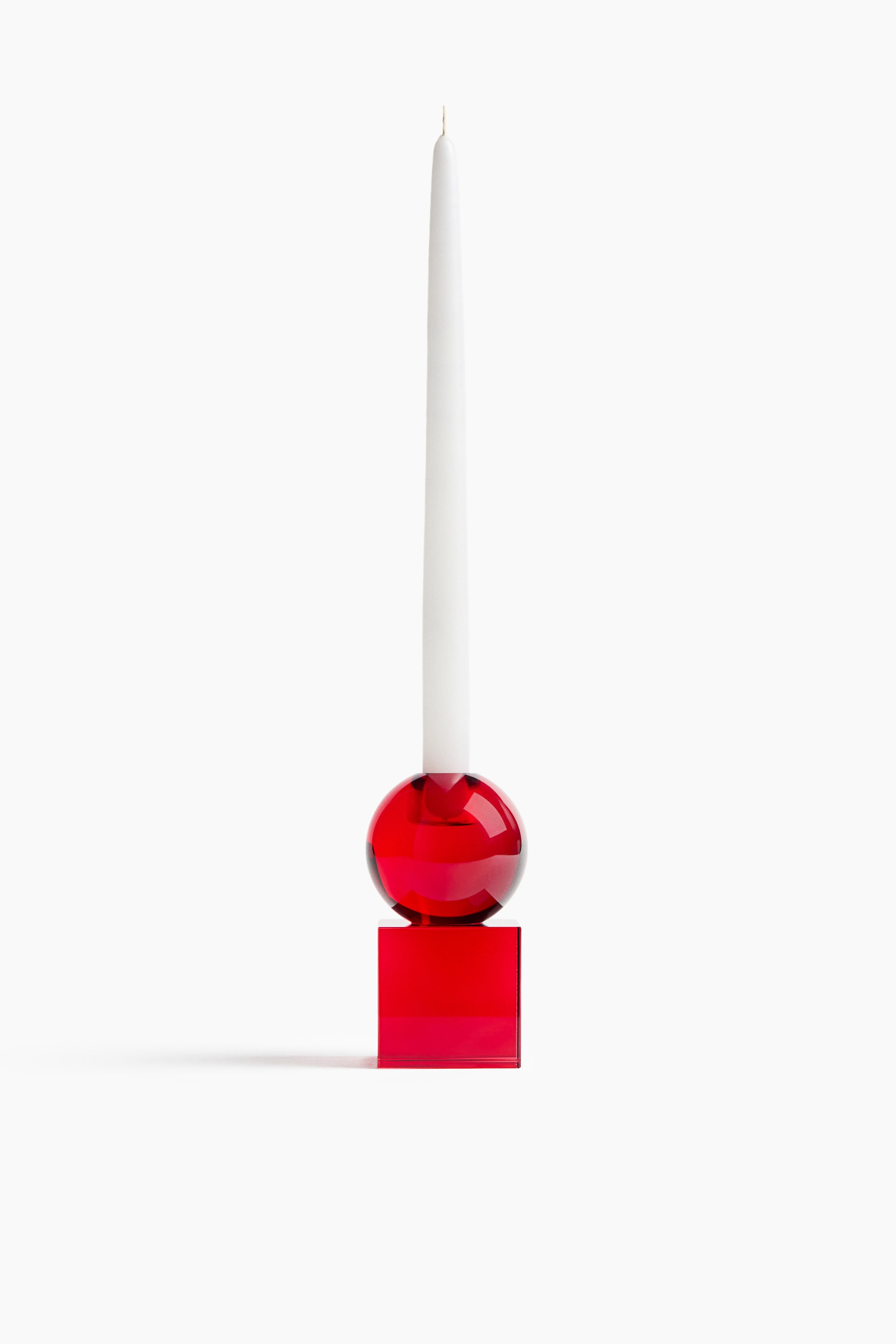 View larger image: The red glass candle holder, shown from the front, features a translucent red cubic base topped with a matching glass sphere. A tall white taper candle stands upright, inserted into the top opening of the sphere.