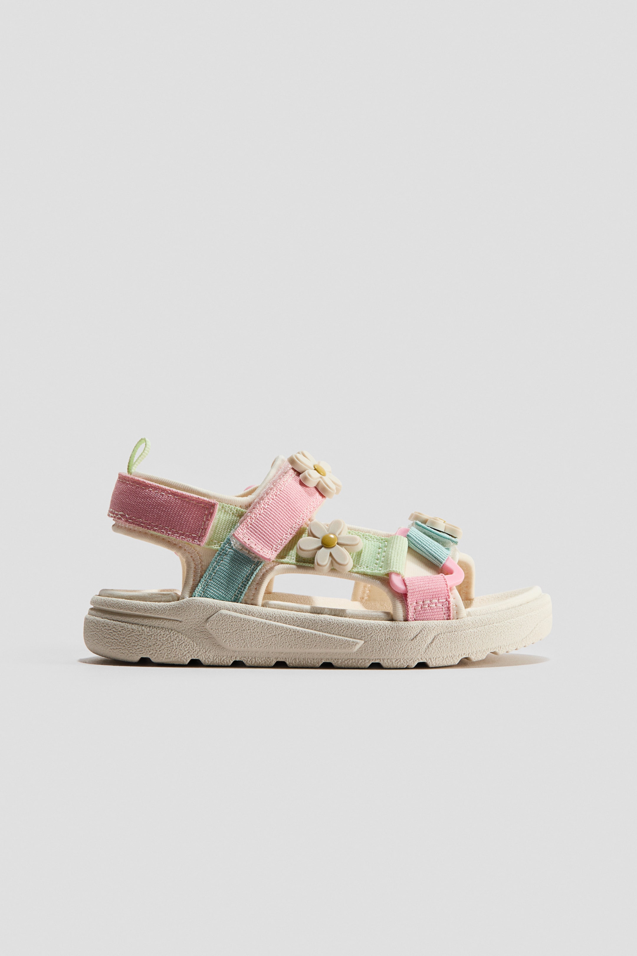 View larger image: A children's sandal is showcasing a side view, featuring multi-colored pastel webbing straps in pink, light green, and light blue crisscrossing across the foot and fastening at the ankle with a hook-and-loop closure. Small, cream-colored flower appliques with yellow centers are decorating the straps, and a chunky, off-white sole with a textured outsole is supporting the sandal.