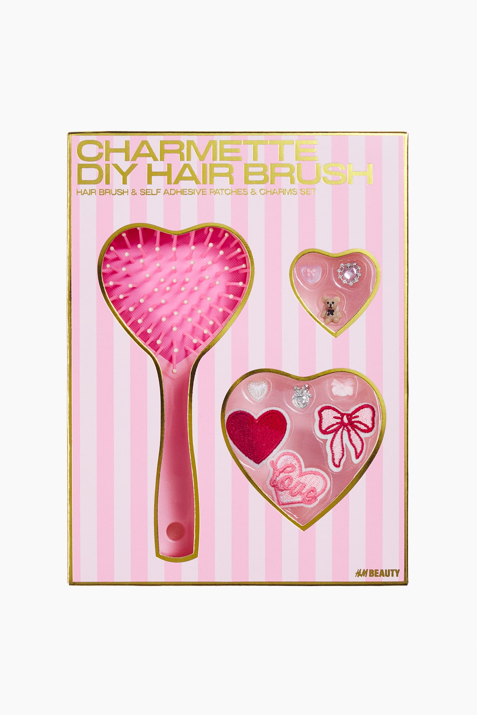 A DIY hairbrush set is shown from the front in a pink-and-white striped box with gold trim. The set features a pink, heart-shaped hairbrush with white-tipped bristles, alongside two heart-shaped compartments containing miniature charms and adhesive patches, including a pink bow and a red heart.