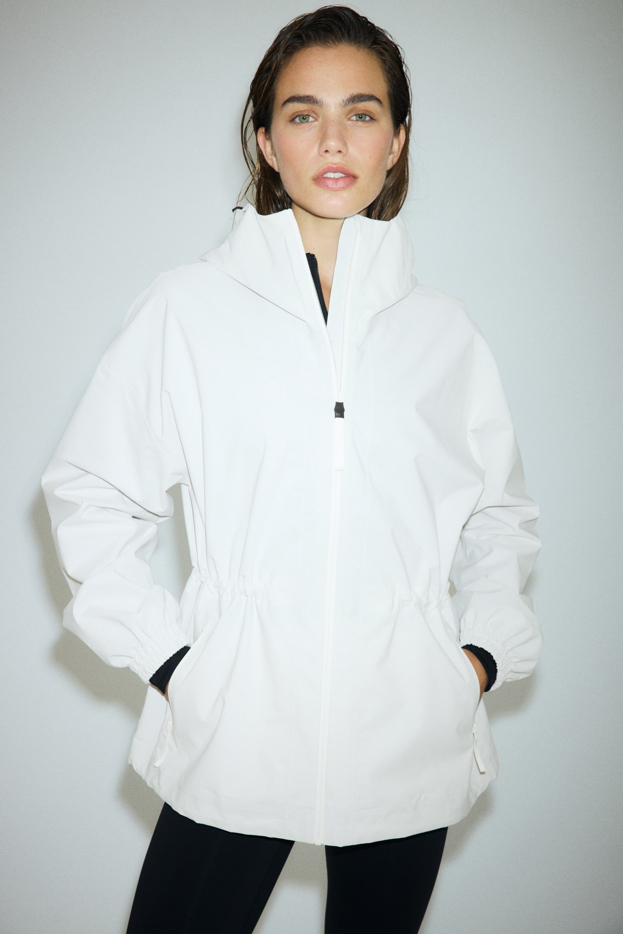 Ver imagen más grande: Una chaqueta anorak blanca está de frente, con un cuello alto con capucha integrada, un cuello interior negro visible y una cremallera frontal con un tirador negro. La chaqueta también muestra una cintura ajustable con cordón que ciñe la prenda, mangas largas que terminan en puños elásticos con puños interiores negros visibles, y dos bolsillos con cremallera en la parte delantera.