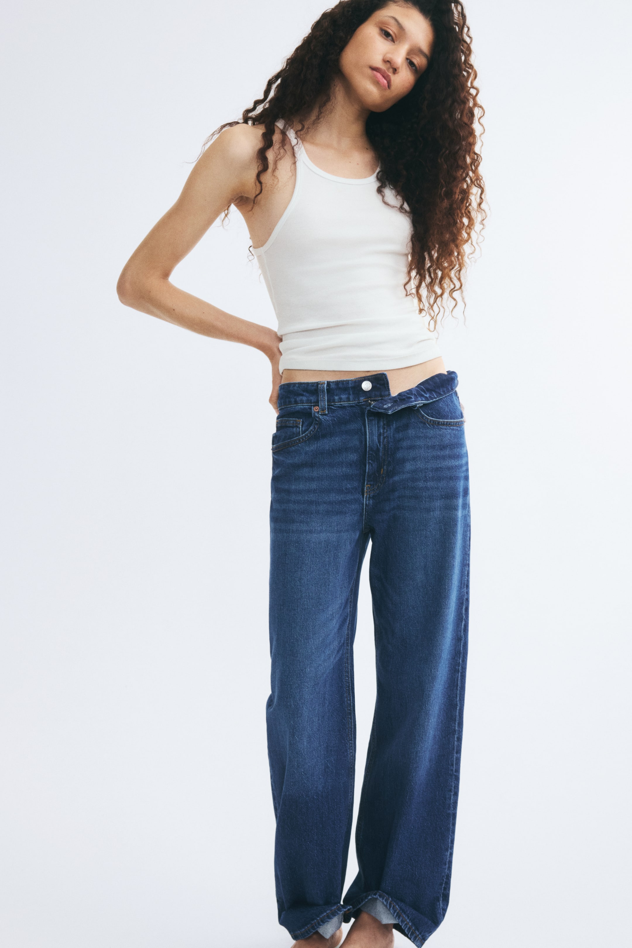 Se større billede: Disse mørkeblå denimjeans med brede ben bæres af modellen og er stylet med en hvid tanktop. Jeansene har en højtaljet fem-lomme-konstruktion, hvor linningen er foldet ned foran for at afsløre en knaplukning.