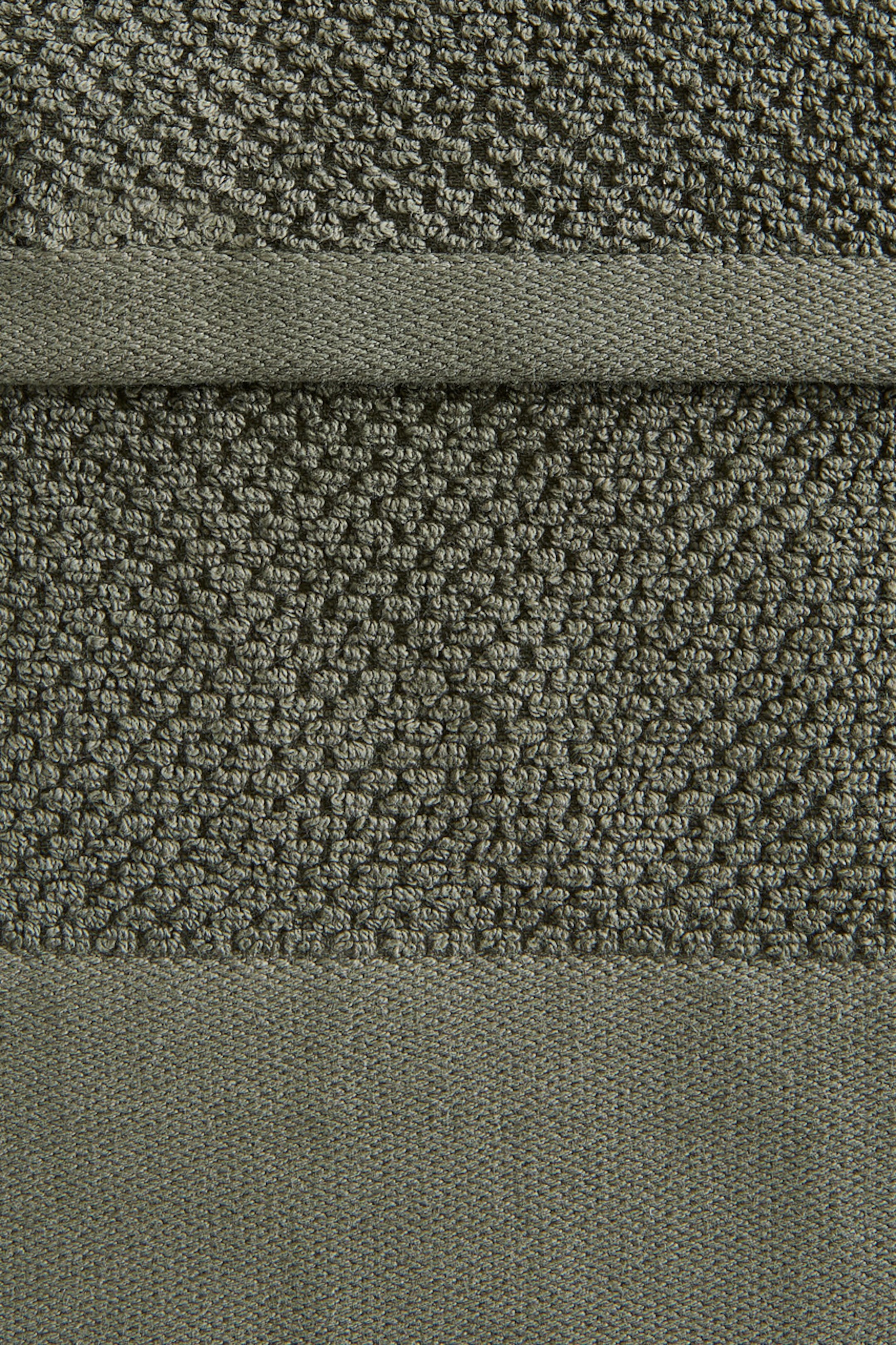 Grösseres Bild ansehen: Ein olivgrüner Stoff zeigt eine strukturierte Oberfläche mit kleinen, erhabenen Schlingen. Er verfügt über zwei horizontal gewebte Bänder, die die Abschnitte der geschlungenen Textur voneinander trennen.