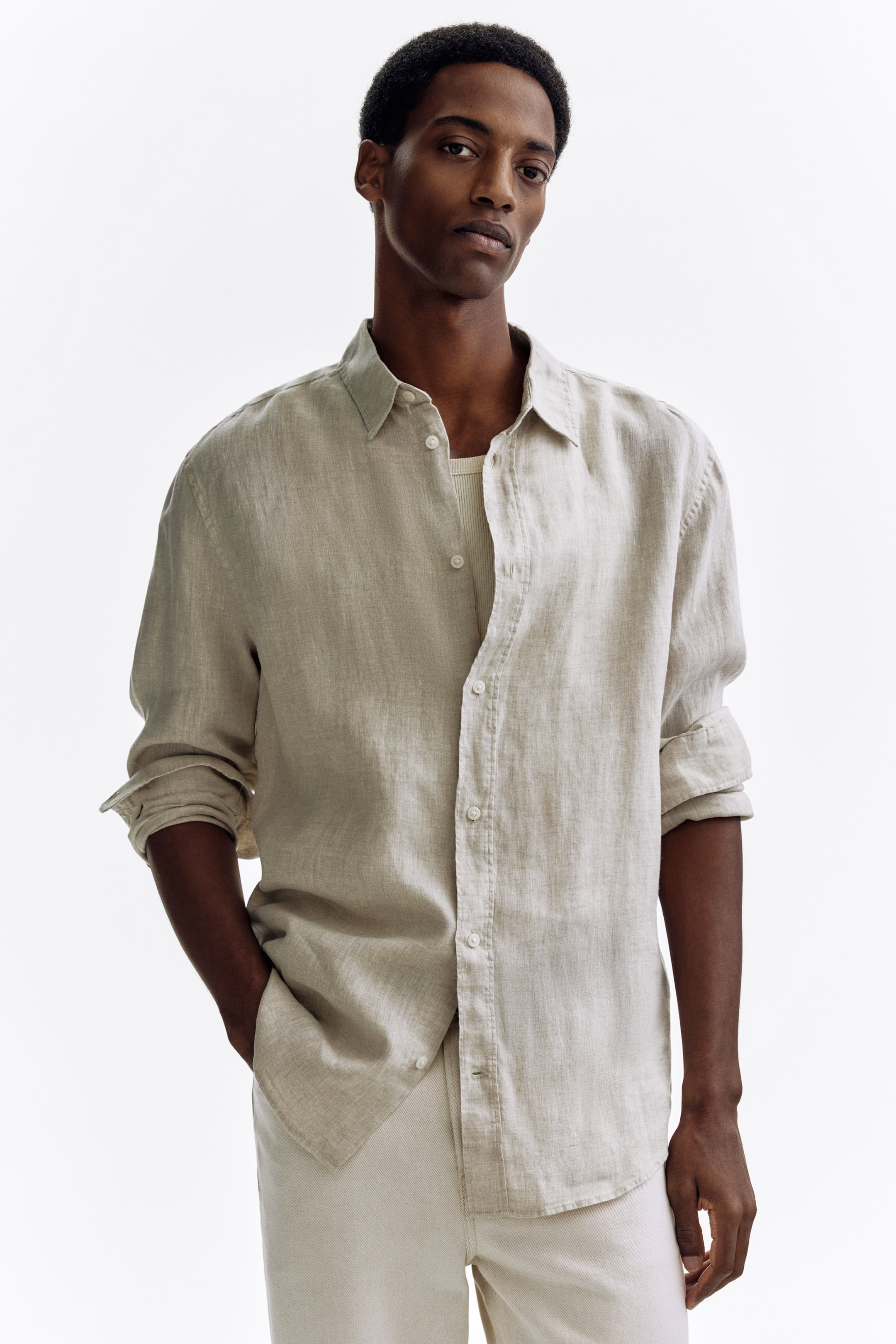 View larger image: A light beige linen shirt with a relaxed fit is being worn, featuring a spread collar and a full button placket with white buttons. Its long sleeves are rolled up, and it is styled unbuttoned over a light ribbed undershirt.