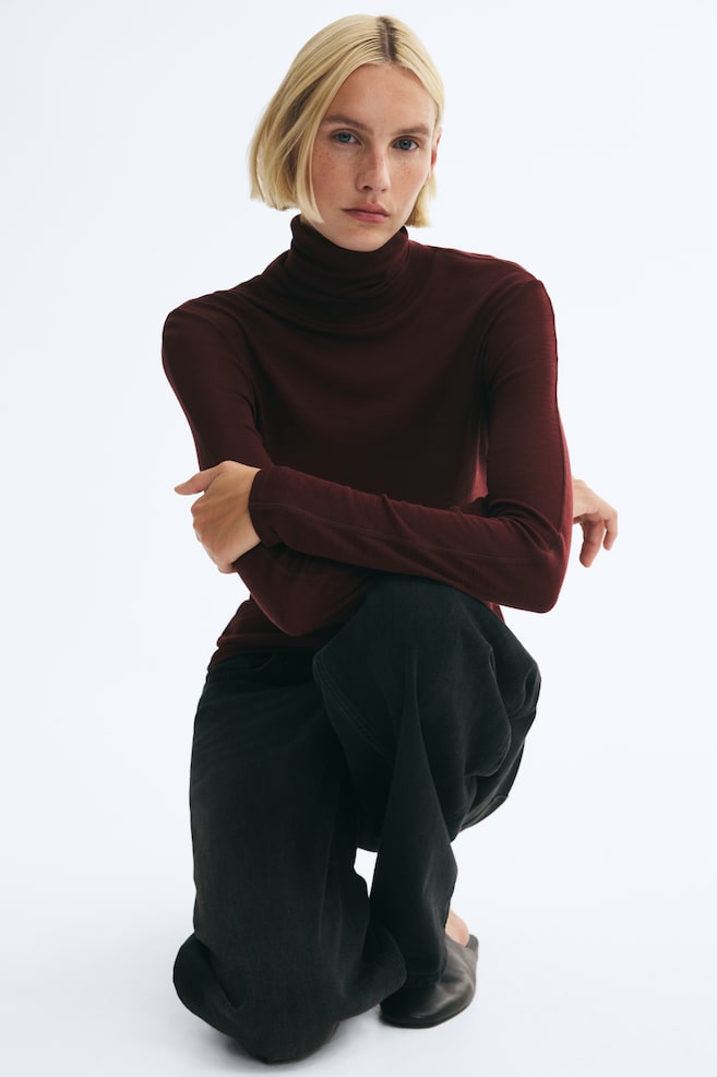 A fitted, dark burgundy turtleneck top in a fine rib knit is worn by the model, who is posing in a crouched position with her arms crossed over one knee. The long-sleeved top is styled with wide-leg black trousers and black leather shoes. - 1