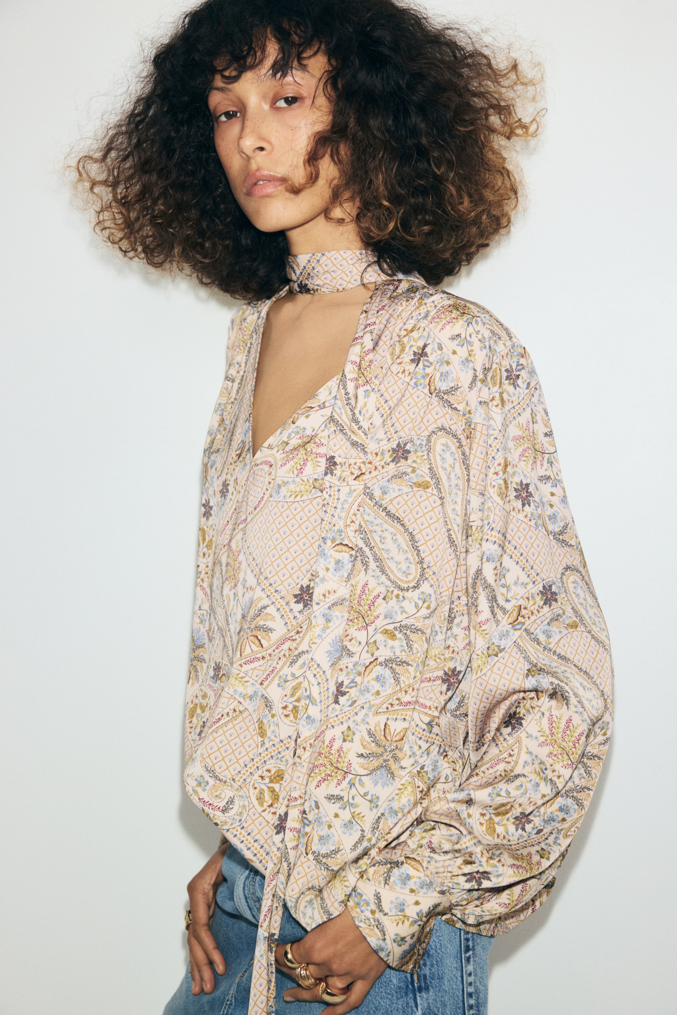 Ampliar la imagen: La modelo lleva puesta una blusa oversize con estampado paisley multicolor. La blusa presenta cuello en V, un cuello tipo pañuelo a juego enrollado alrededor del cuello y mangas largas fruncidas, y se combina con jeans azules.