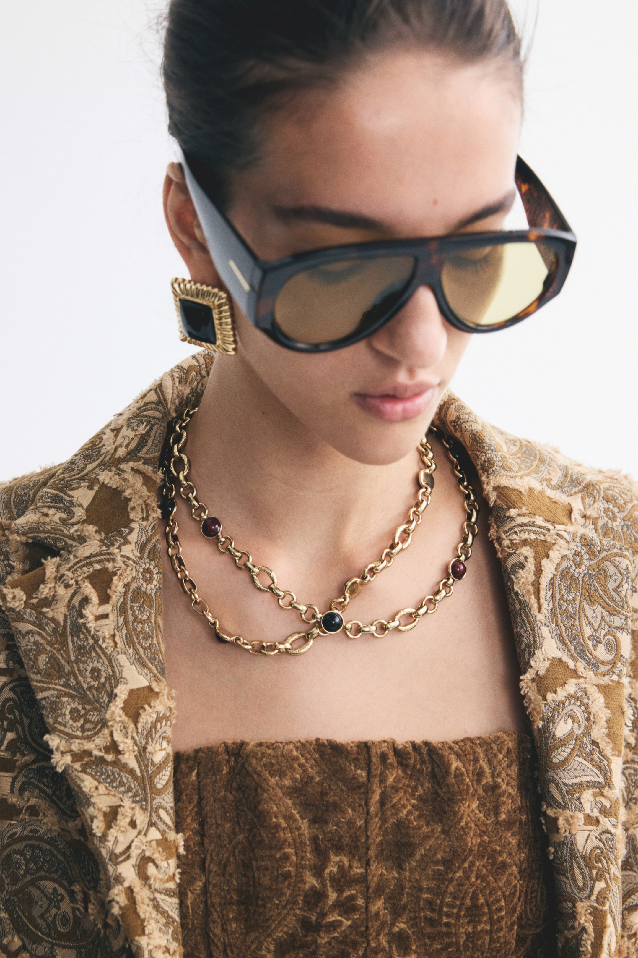Visualizza l'immagine ingrandita: La modella indossa una collana a catena dorata stratificata con piccoli inserti in pietra rotondi, abbinata a grandi orecchini squadrati in oro con texture e centro nero. Gli accessori sono completati da occhiali da sole oversize effetto tartaruga con lenti sfumate gialle e una giacca marrone paisley con texture.