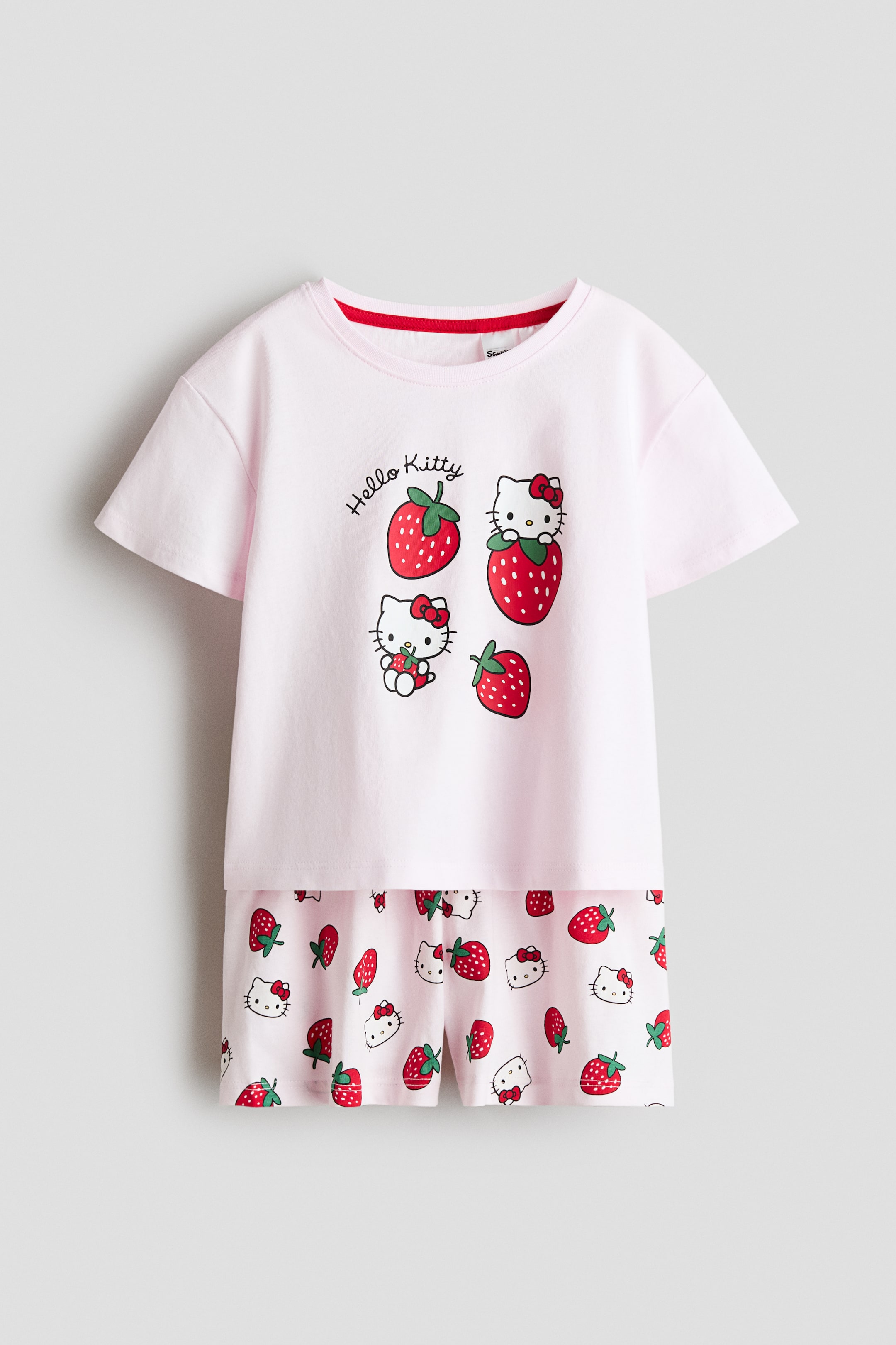 Näytä suurempi kuva: Kaksiosainen yöasusetti esittelee etunäkymänsä. Vaaleanpunaisessa t-paidassa on lyhyet hihat, pyöreä pääntie punaisella sisäkantilla ja etupuolella graafinen printti, jossa on "Hello Kitty" -teksti, erilaisia Hello Kitty -hahmoja ja mansikoita. Yhteensopivat vaaleanpunaiset shortsit esittelevät kauttaaltaan kuvioinnin, jossa on Hello Kitty -päitä ja mansikoita.