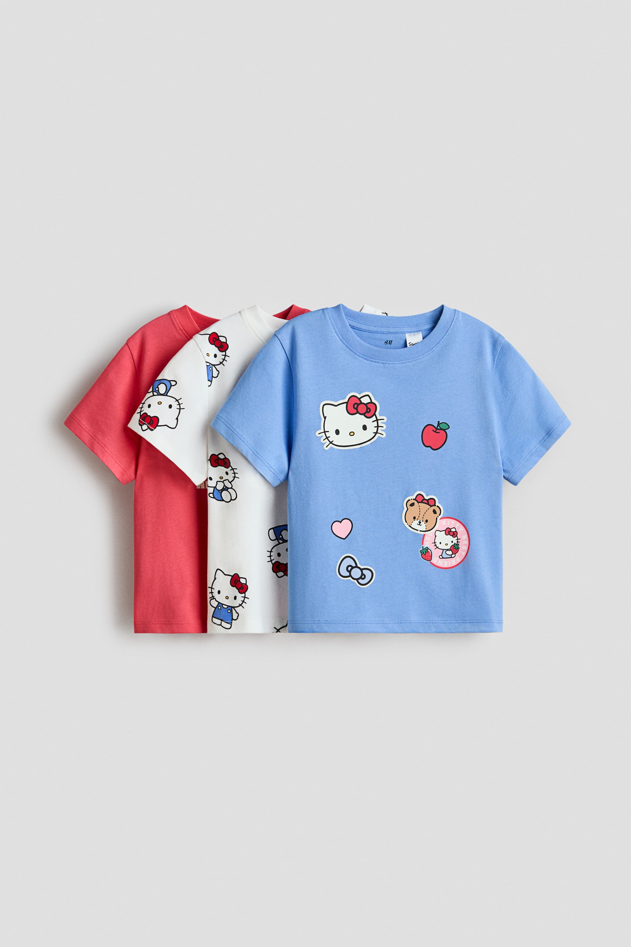 Voir une image plus grande: Trois hauts en jersey de coton à manches courtes sont présentés en superposition, face avant. Le haut bleu à l'avant arbore divers motifs de Hello Kitty et de personnages, y compris une pomme et un cœur. Les hauts blanc et corail derrière affichent des motifs Hello Kitty sur toute la surface.