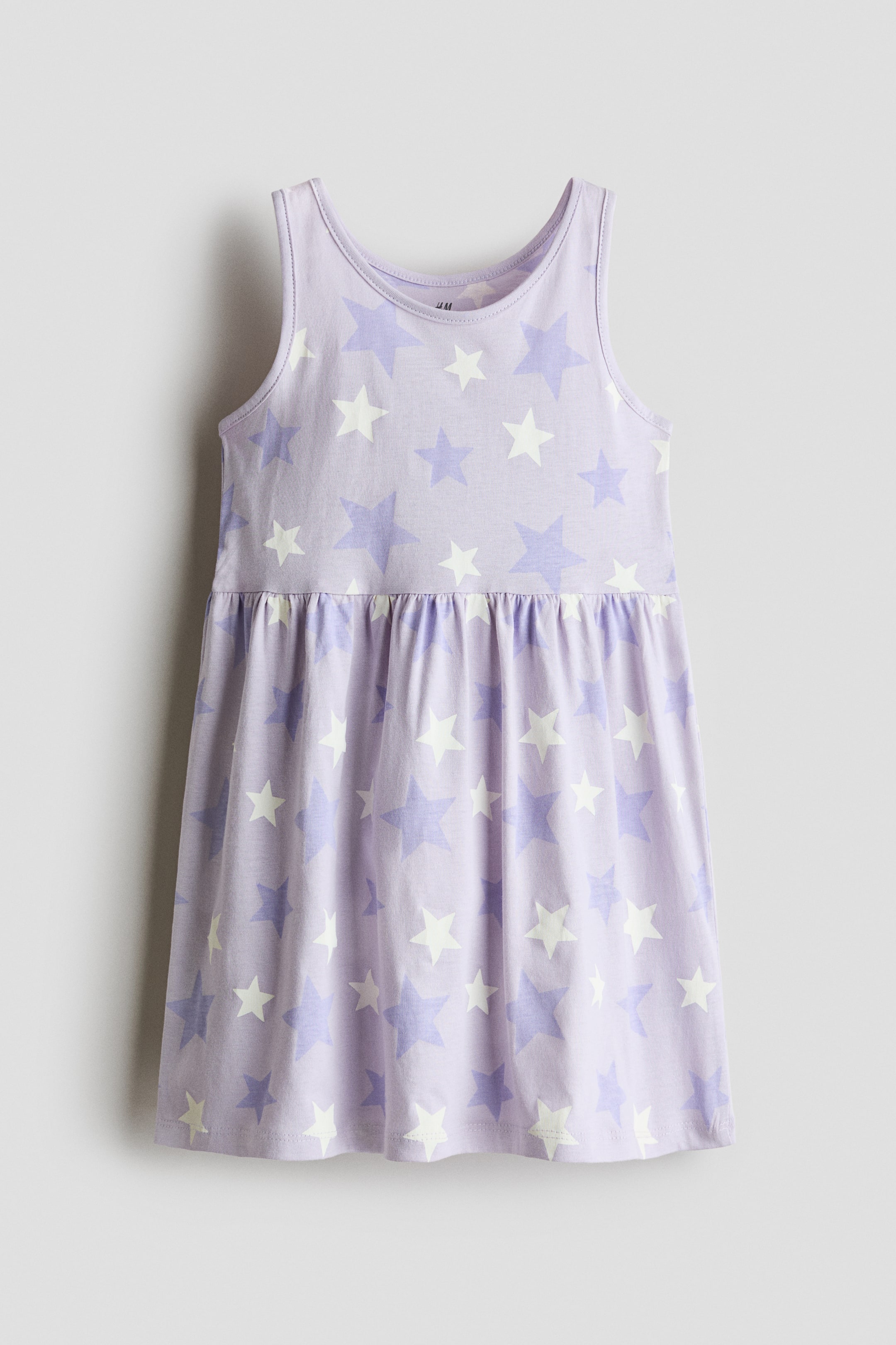 View larger image: A light purple sleeveless dress, front-facing, features an all-over print of scattered white and medium-purple stars. The dress has a scoop neckline, wide shoulder straps, and a gathered waist that flows into a flared skirt.