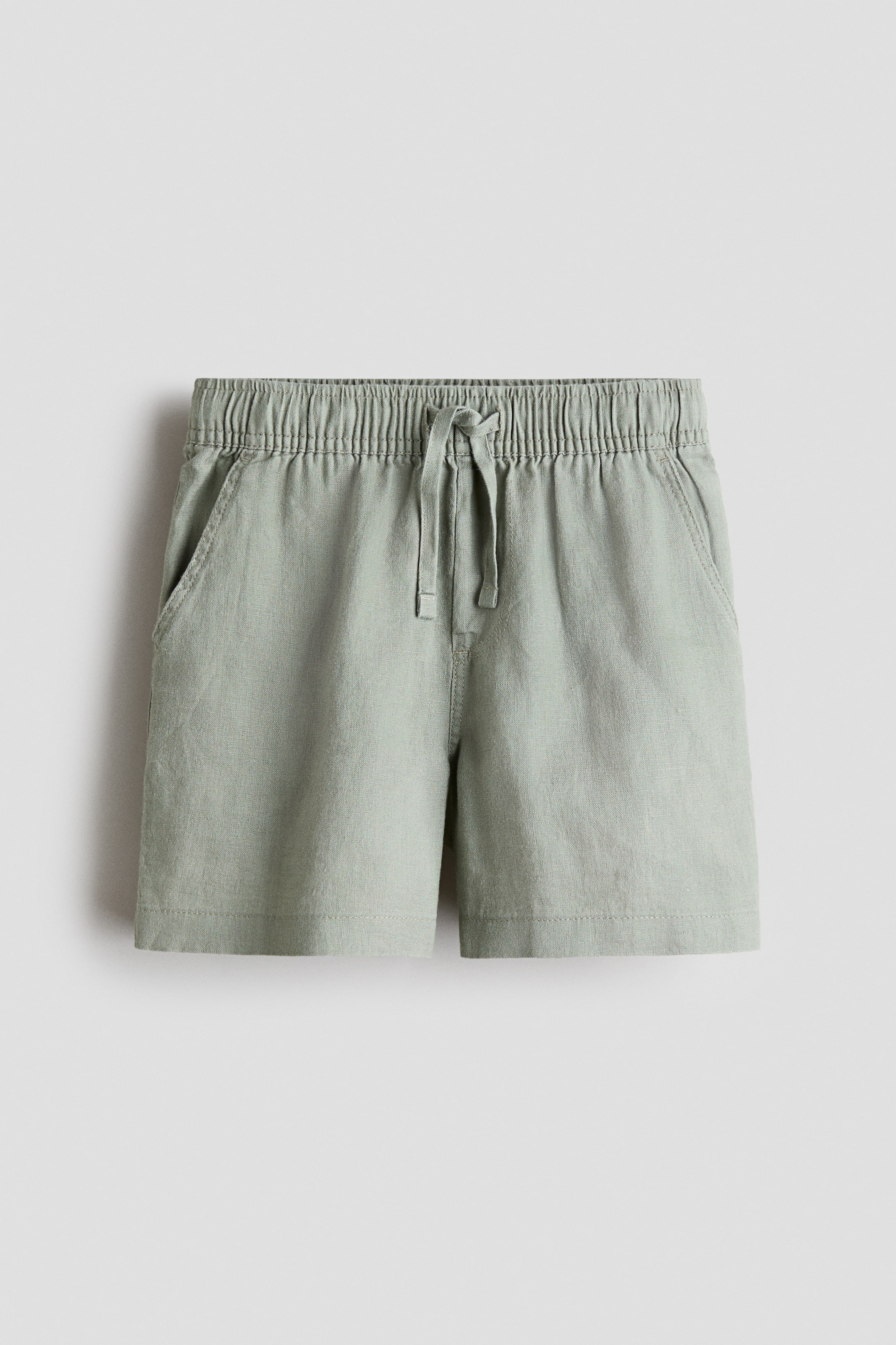 View larger image: A pair of light green linen shorts is showcased from the front, featuring an elasticated, ruched waistband with a tied drawstring, a faux fly, and diagonal side pockets.