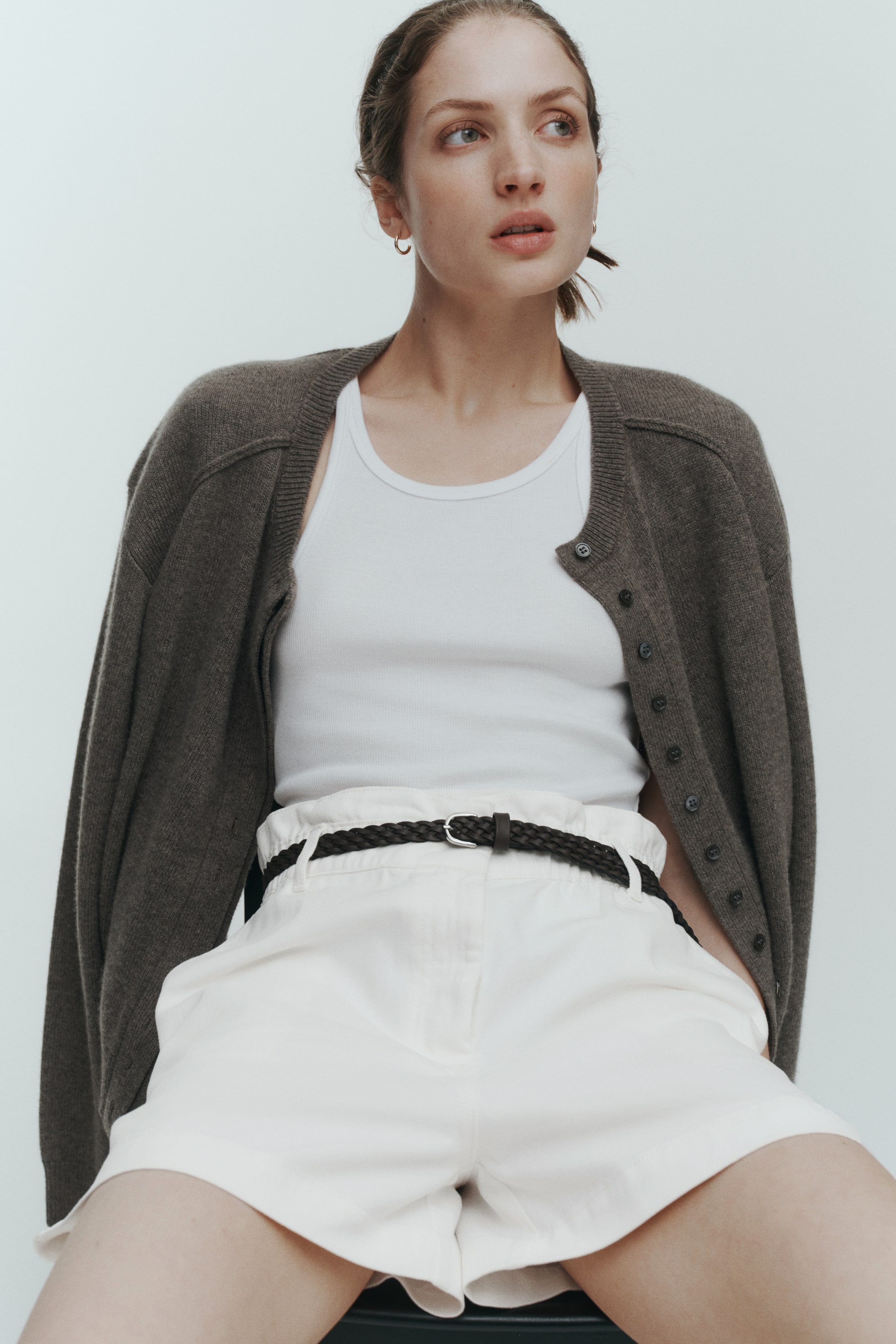 View larger image: Loose-fit shorts in light-coloured cotton twill are worn by the model, featuring a high waist with gathered elastication, a button fastening, and diagonal front pockets. A narrow, dark braided belt with a metal buckle cinches the waist of the shorts.