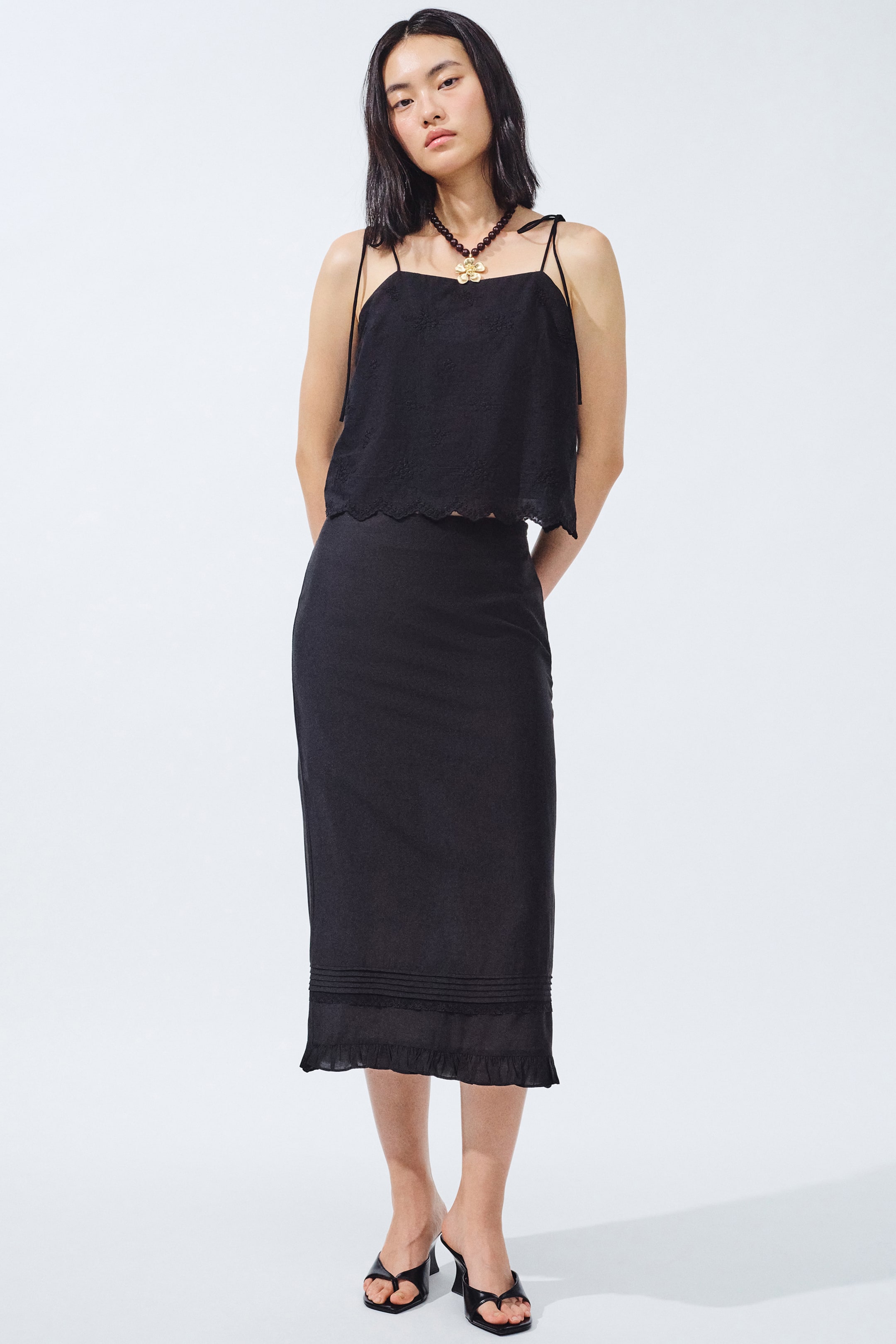 View larger image: The black two-piece ensemble is showcased from the front. The camisole top boasts tied spaghetti straps, a straight neckline, and a scalloped hem with delicate embroidered accents. The fitted midi skirt features horizontal pintucks above a ruffled hem.