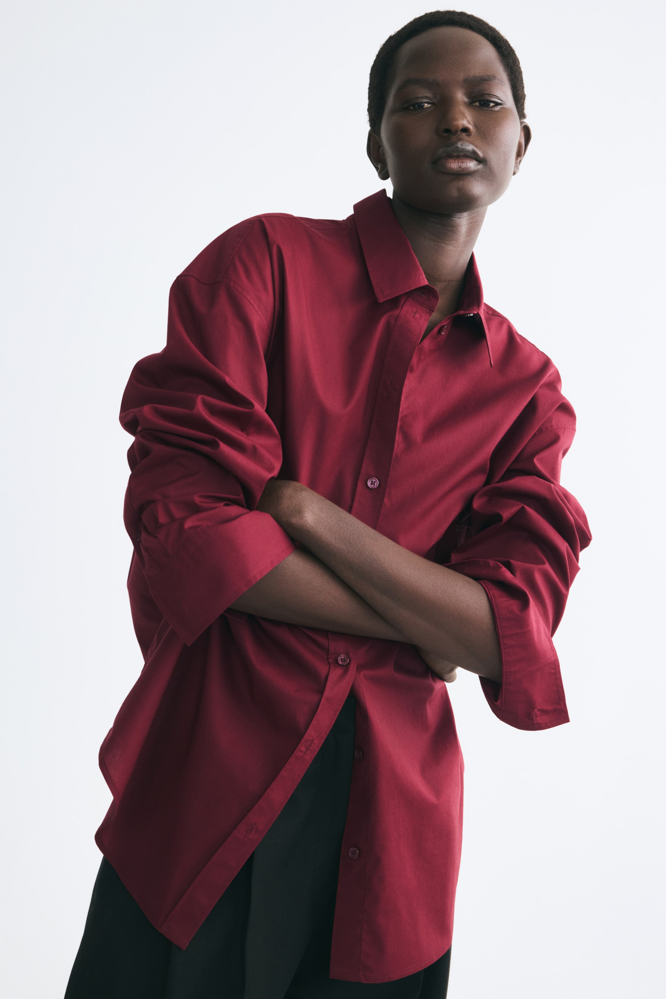 Grotere afbeelding bekijken: Het model draagt een oversized, donkerrode geweven blouse en staat met de armen over elkaar. De blouse heeft een puntkraag, een knoopsluiting aan de voorkant en lange mouwen die tot aan de ellebogen zijn opgestroopt met omgeslagen manchetten, en is aan de onderkant gedeeltelijk losgeknoopt over een zwarte broek.